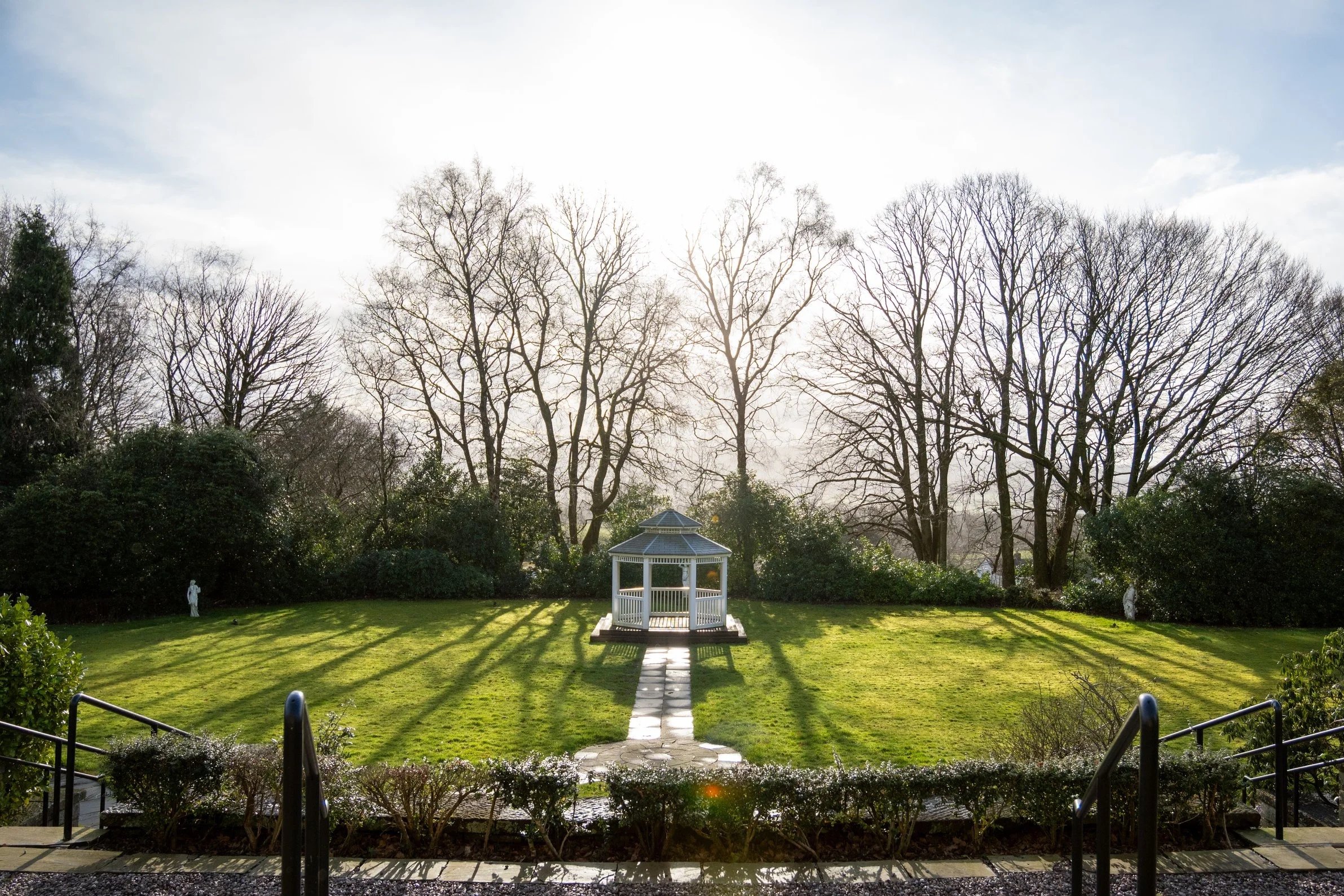 A small white gazebo on a grassy lawn with trees in the background and a walkway leading up to it, taken during the daytime with sunlight casting shadows.