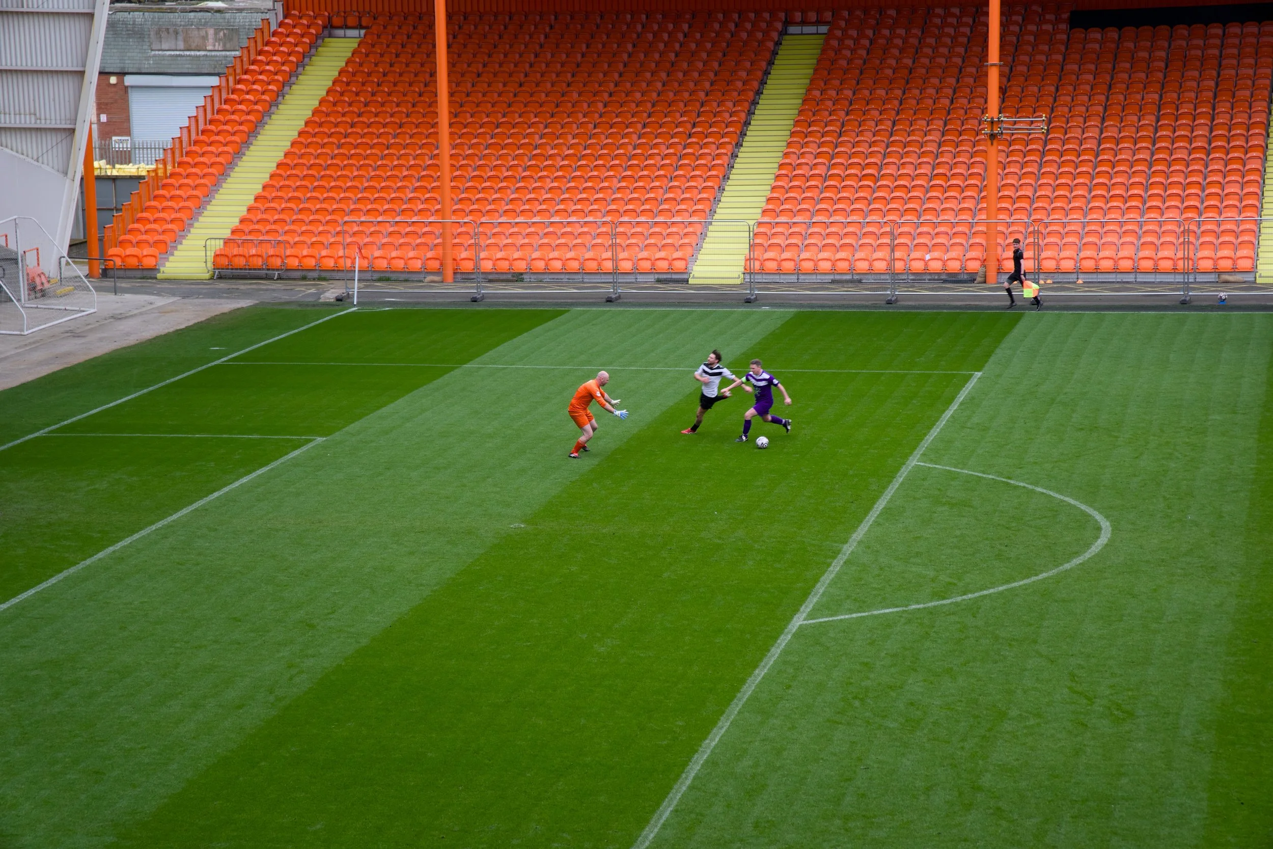 A soccer stadium with orange seats, green stairs, and a few people on the field, including a goalkeeper, players, and a person with a cone.