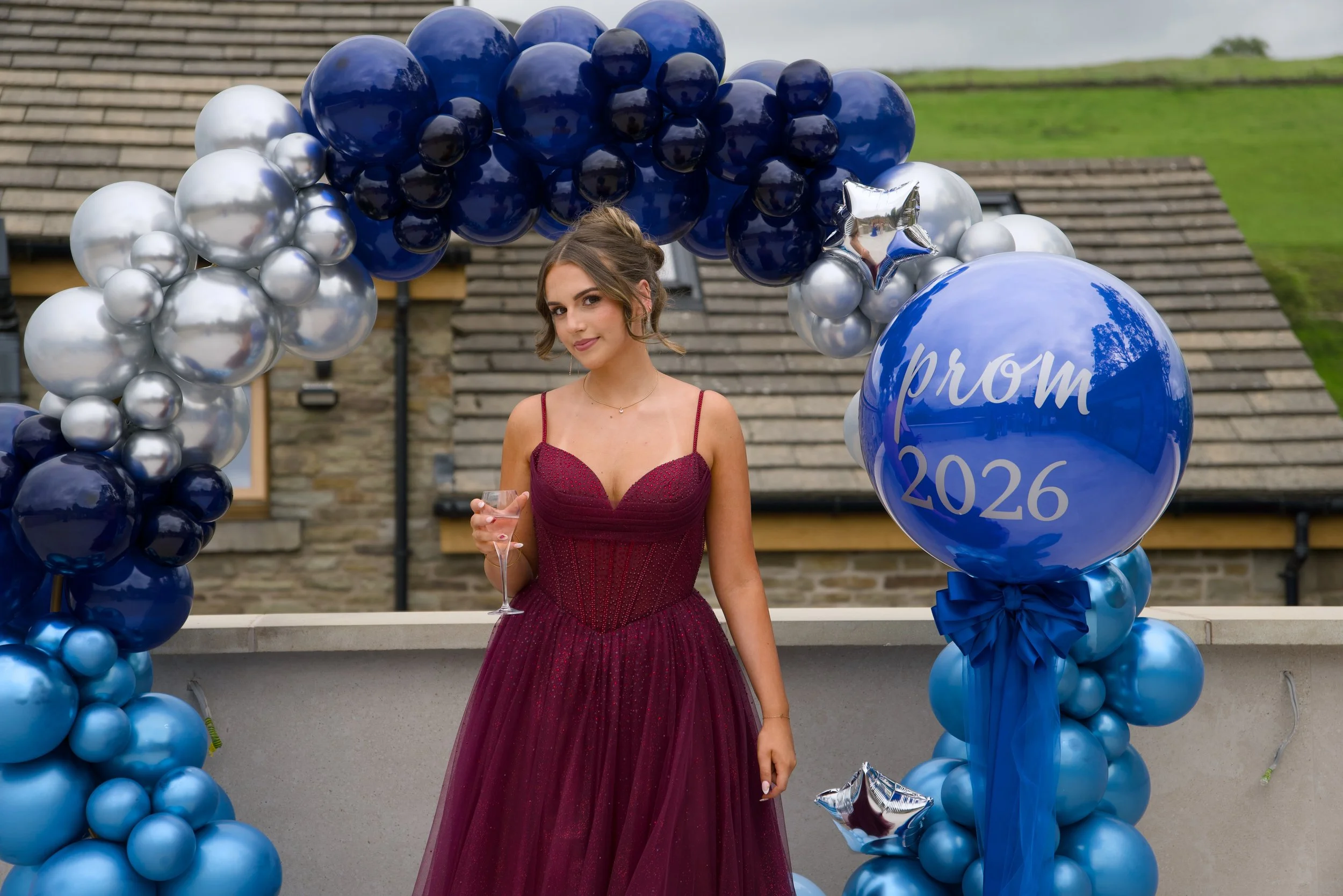 A young woman in a burgundy formal gown stands outdoors in front of a balloon arch. The arch is decorated with blue, silver, and dark blue balloons, with a large blue balloon on the right side displaying white text that reads 'prom 2026.' The woman has brown hair styled in an updo, is holding a champagne flute, and is smiling lightly while looking at the camera.