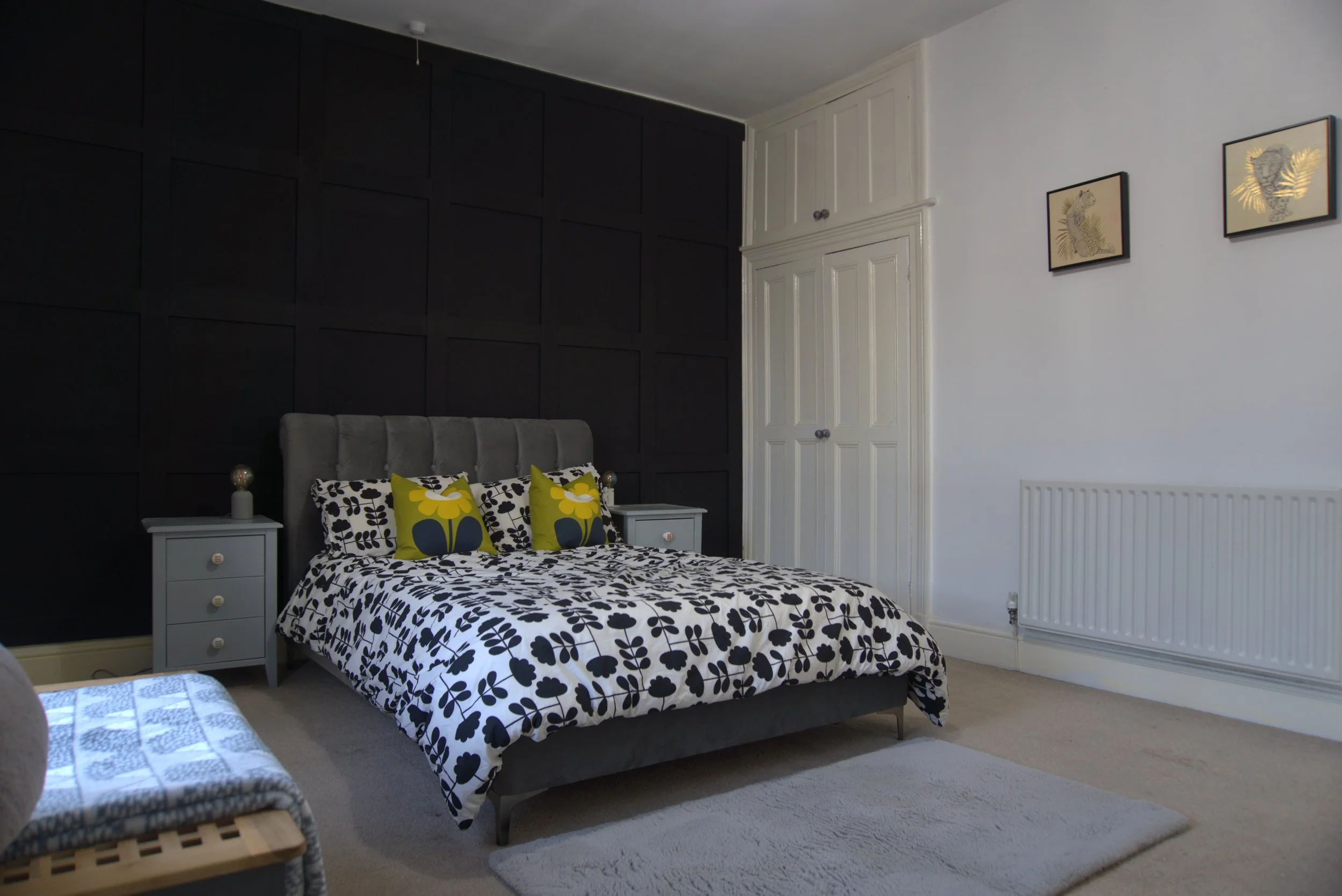 A bedroom with a bed featuring black and white floral bedding, two yellow and black pillows, a gray upholstered headboard, white nightstands with small lamps, a dark accent wall, white built-in cupboard doors, framed artwork of animals on the wall, a white radiator, and a beige carpet.
