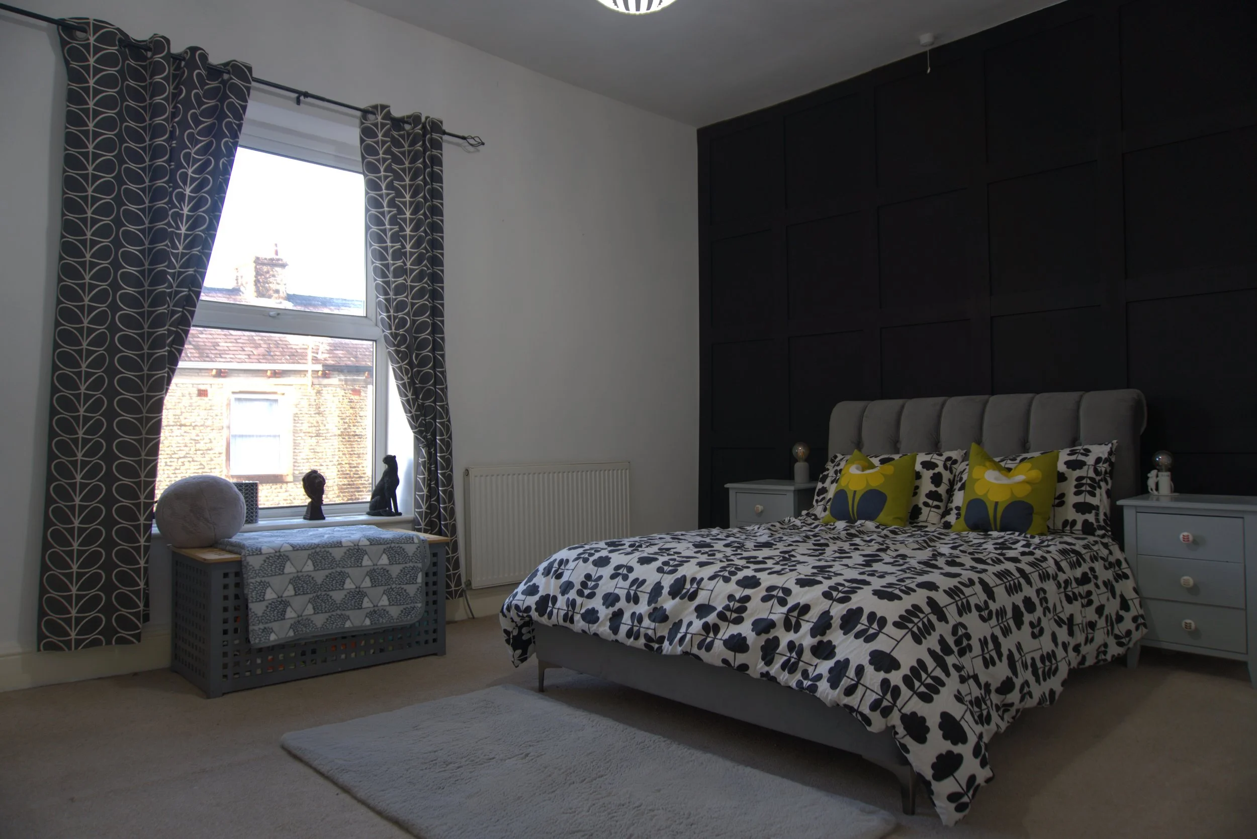A bedroom with a large window covered by dark patterned curtains, a white bed with black and yellow floral bedding, white bedside tables, and various decorative items on a window sill, including sculptures and a patterned bench with a round cushion.