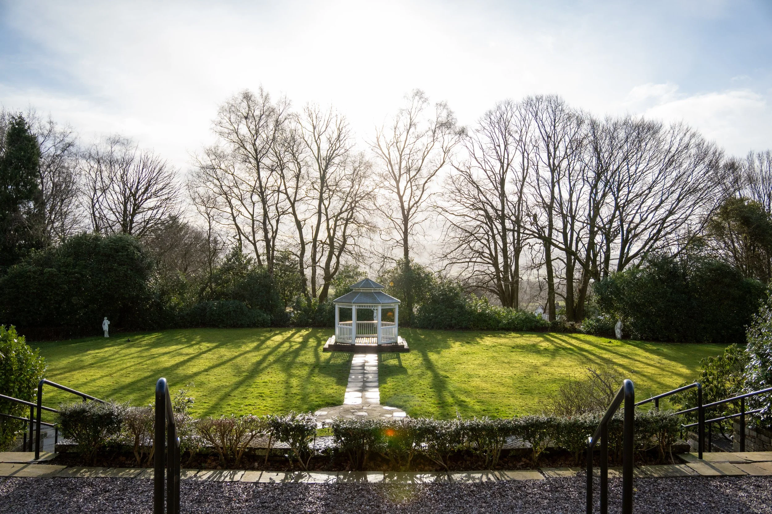 Sunlight shining on a green lawn with a small white gazebo in the center, surrounded by leafless trees and bushes. Shadowed railing and steps in the foreground.