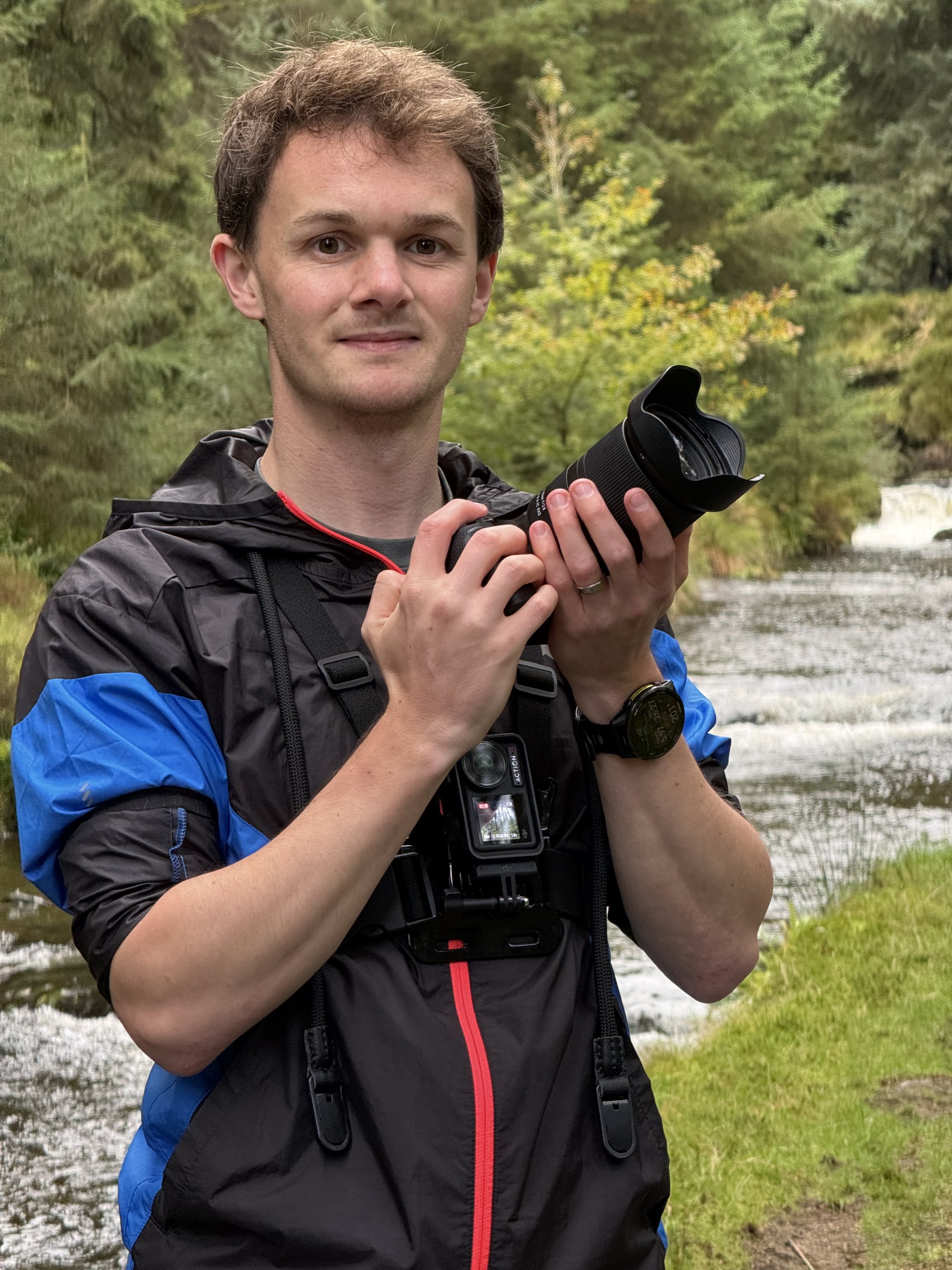 A young man holding a camera outdoors near a river with lush green trees in the background.