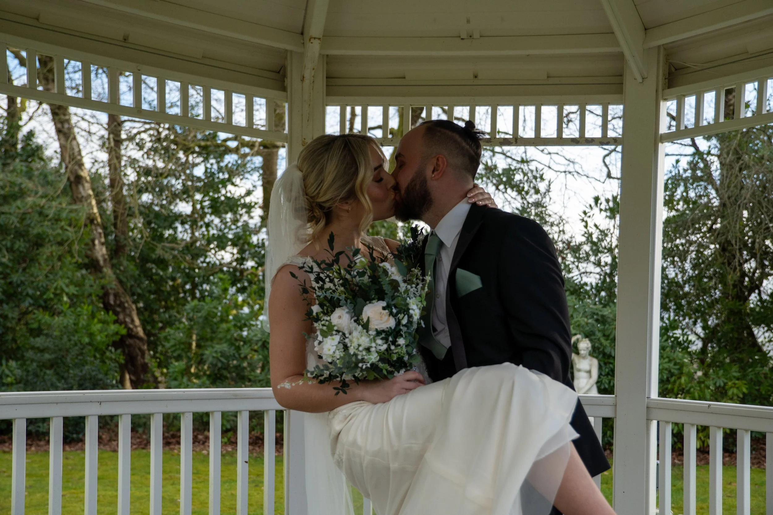 A bride and groom sharing a kiss on a gazebo with trees in the background, the bride holding a bouquet of flowers.