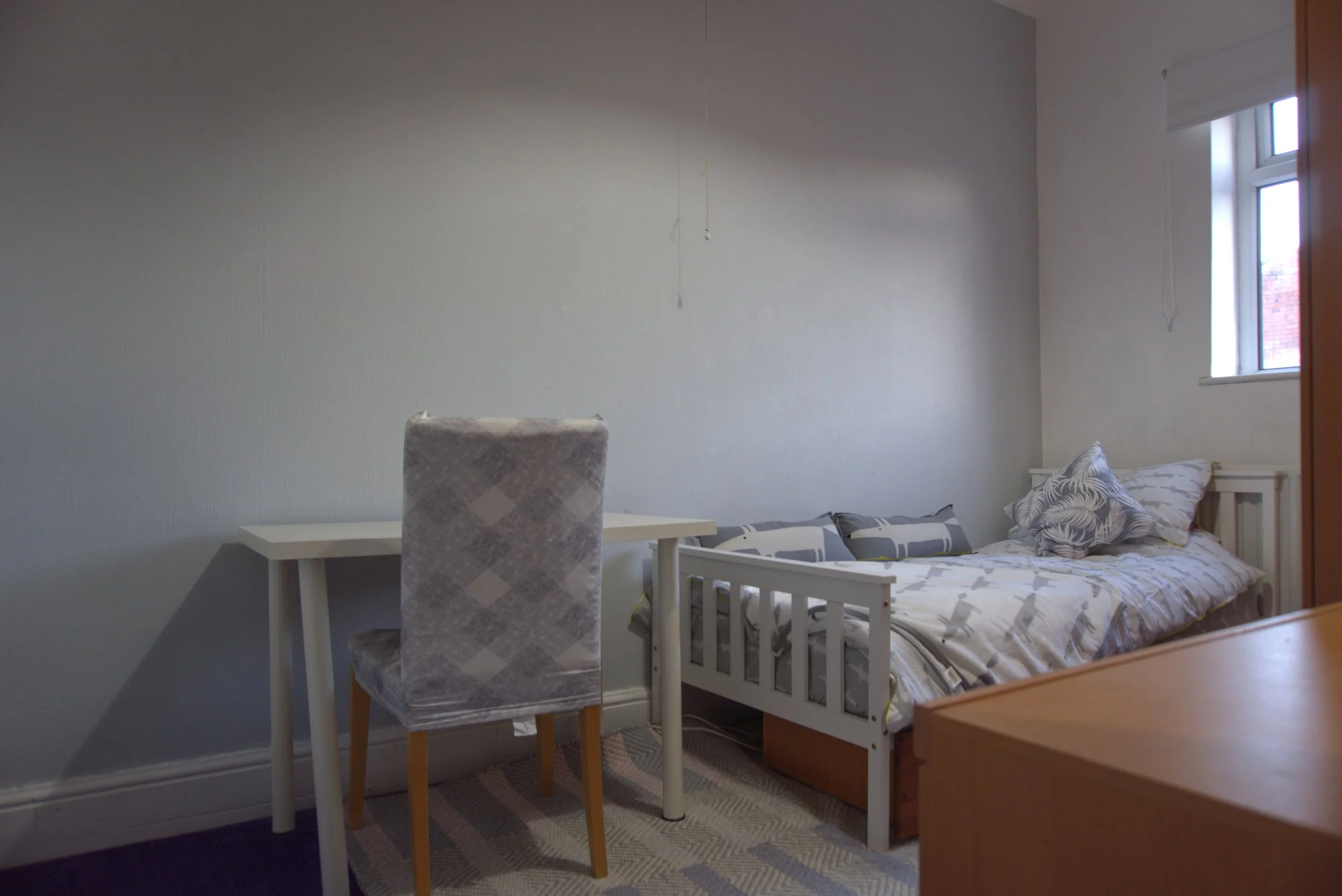 A bedroom with a white bed, pillows, and a blanket, placed next to a window. There is a wooden dresser and a small white table with a patterned chair. The walls are painted light gray, and there is a beige and gray patterned rug on the floor.