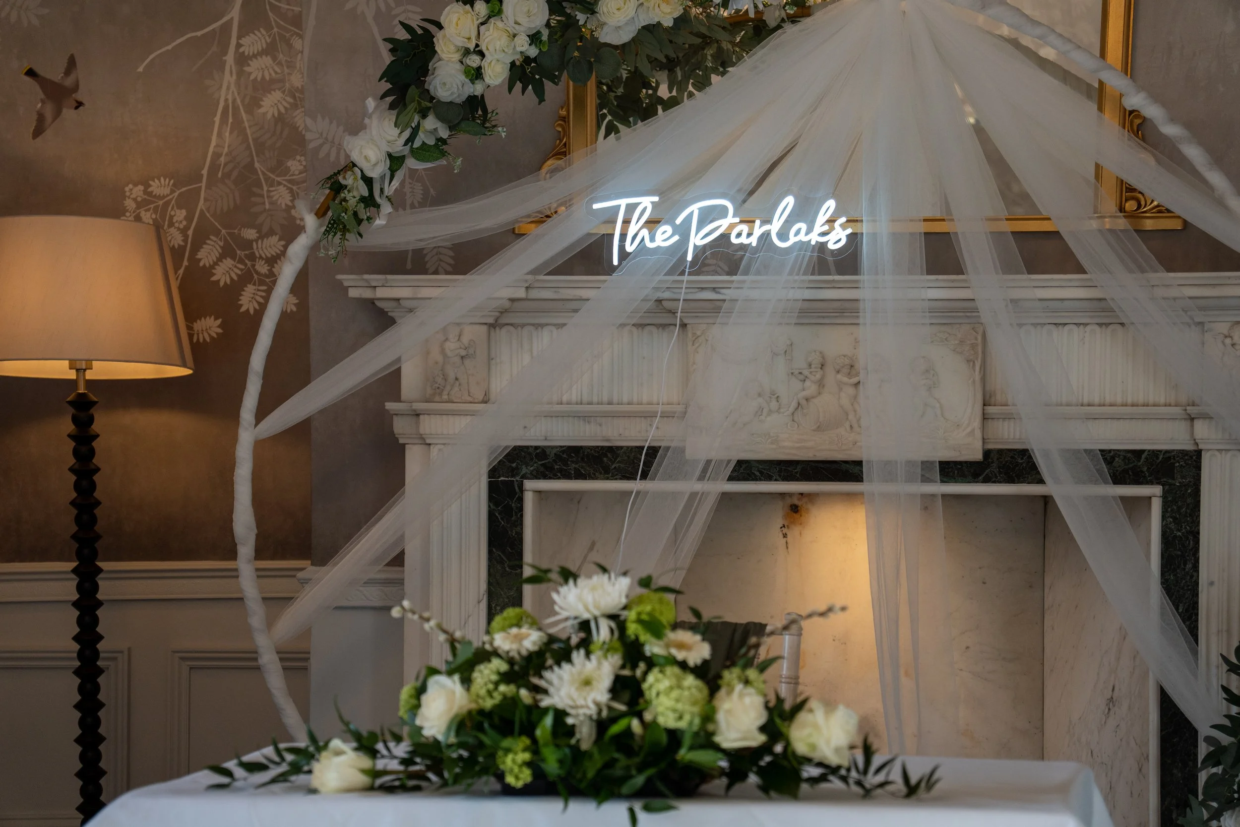 Wedding decor with a white floral arrangement, sheer drapes, and a neon sign that reads 'The Parlaks' in front of a fireplace.