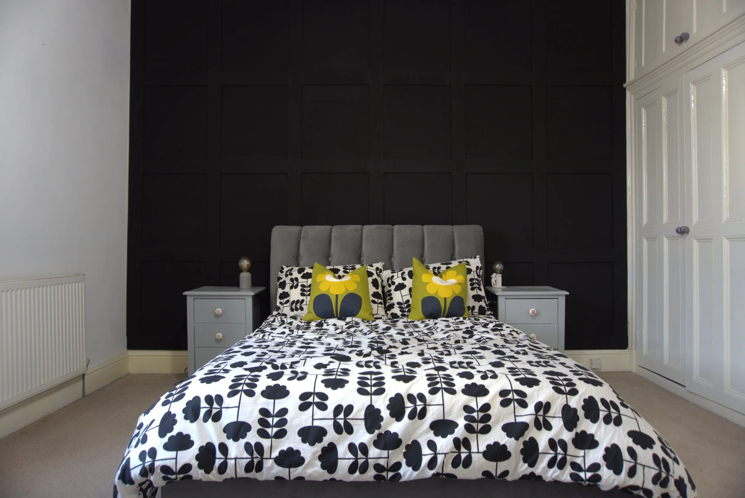 A bed with black and white floral bedding and two pillows with a yellow flower design. The bed is flanked by white nightstands and set against a dark paneled accent wall in a bedroom.