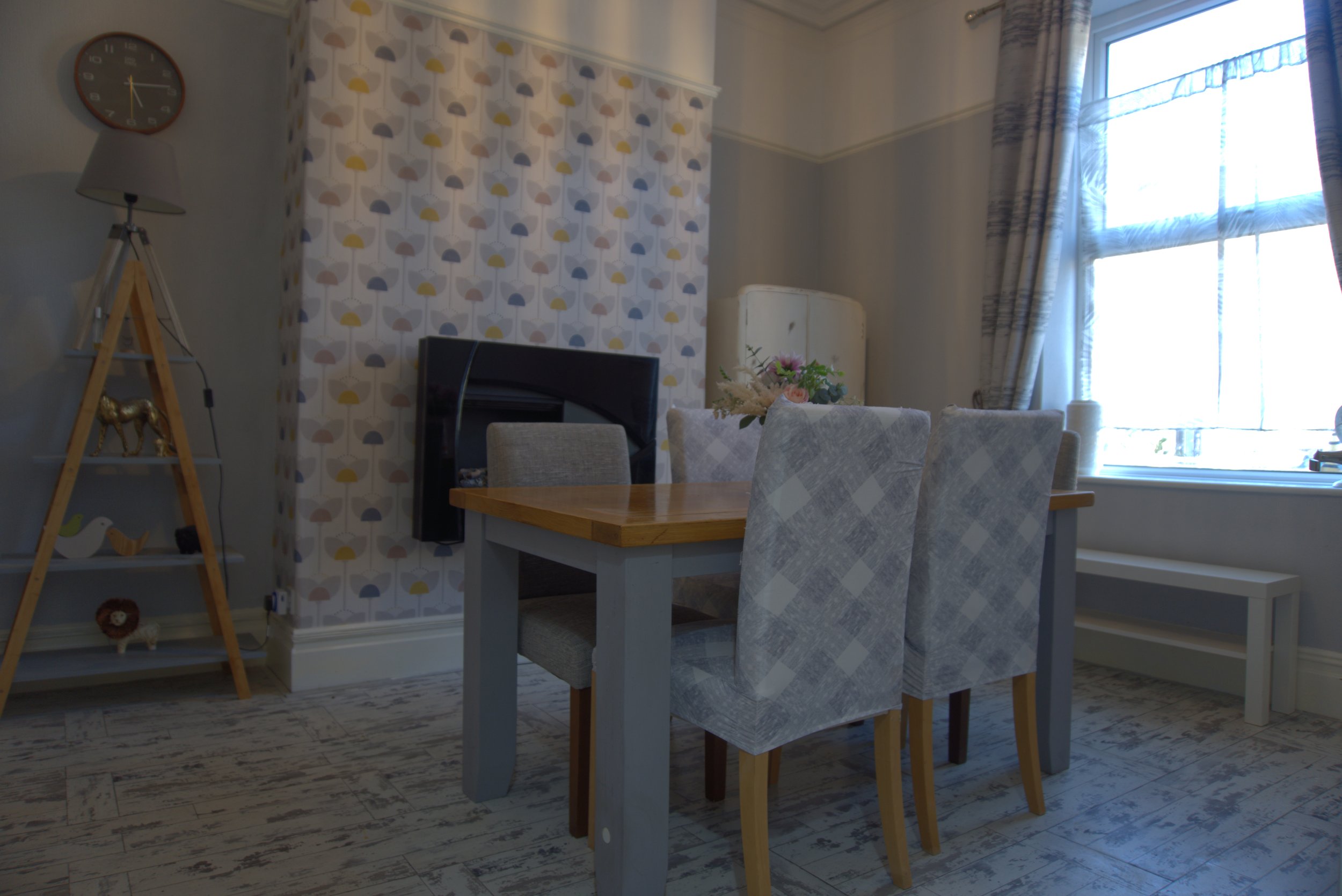 A dining room with a wooden table surrounded by four chairs with patterned fabric. There is a floral centerpiece on the table. A window with curtains lets in natural light. To the left, a shelf with decorative items including a golden horse figurine 