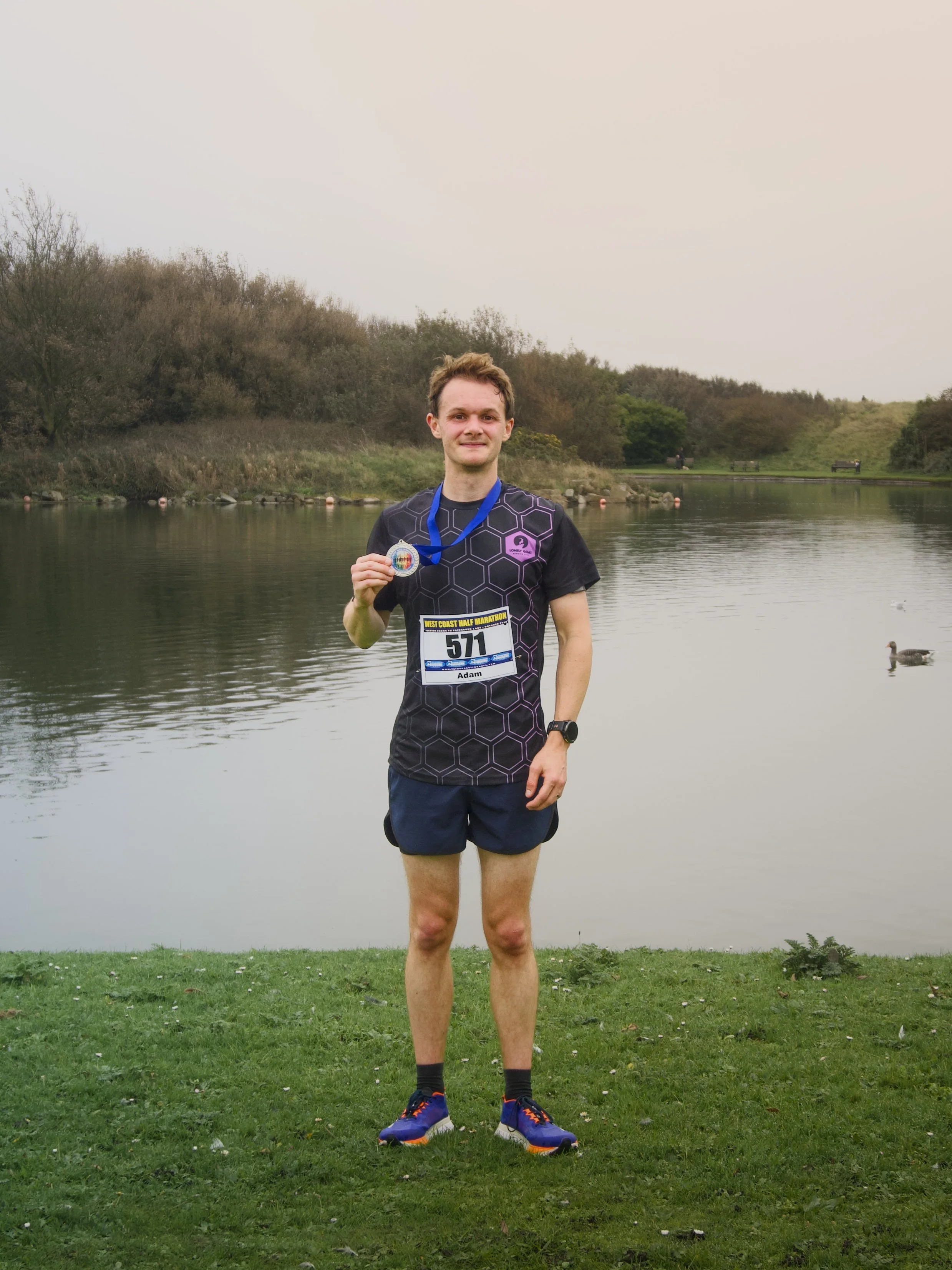 Young man standing by a lake holding a medal, wearing running gear, with trees and benches in the background.