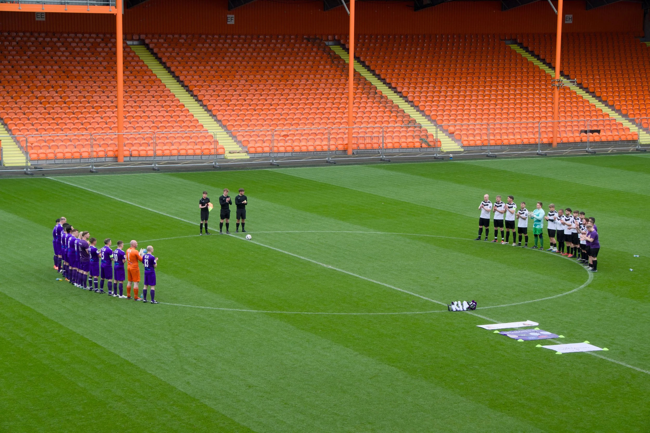 A soccer field with two teams of players and referees standing in line for a pre-game or halftime ceremony. The stands are mostly empty with orange seats.
