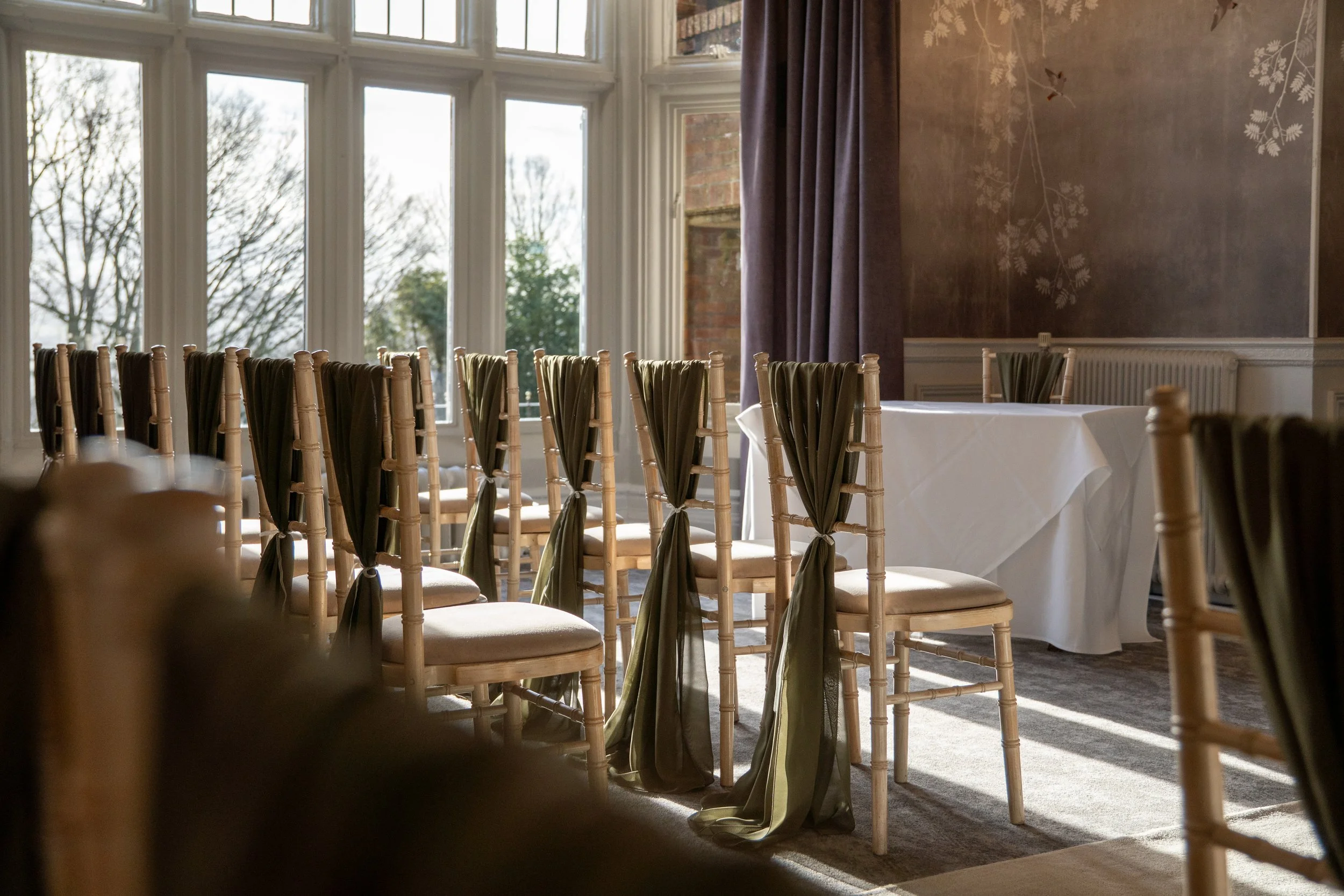 Empty chairs with green sashes in a sunlit, elegant dining room with large windows and white tablecloth-covered tables.
