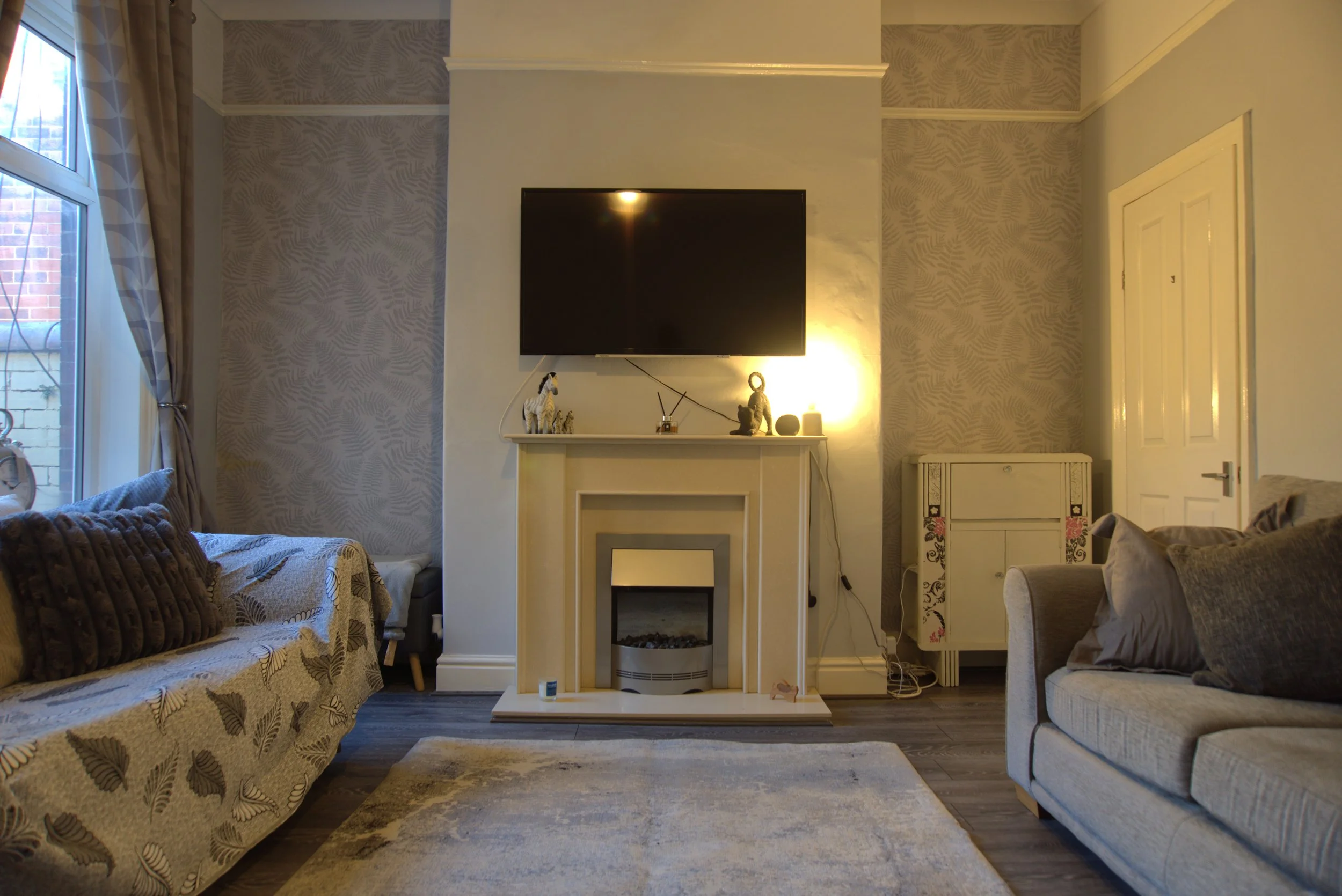 Living room with a TV above a fireplace, beige walls with patterned wallpaper, two sofas with cushions, a rug, and decorative items on the mantle. A window with curtains is on the left.