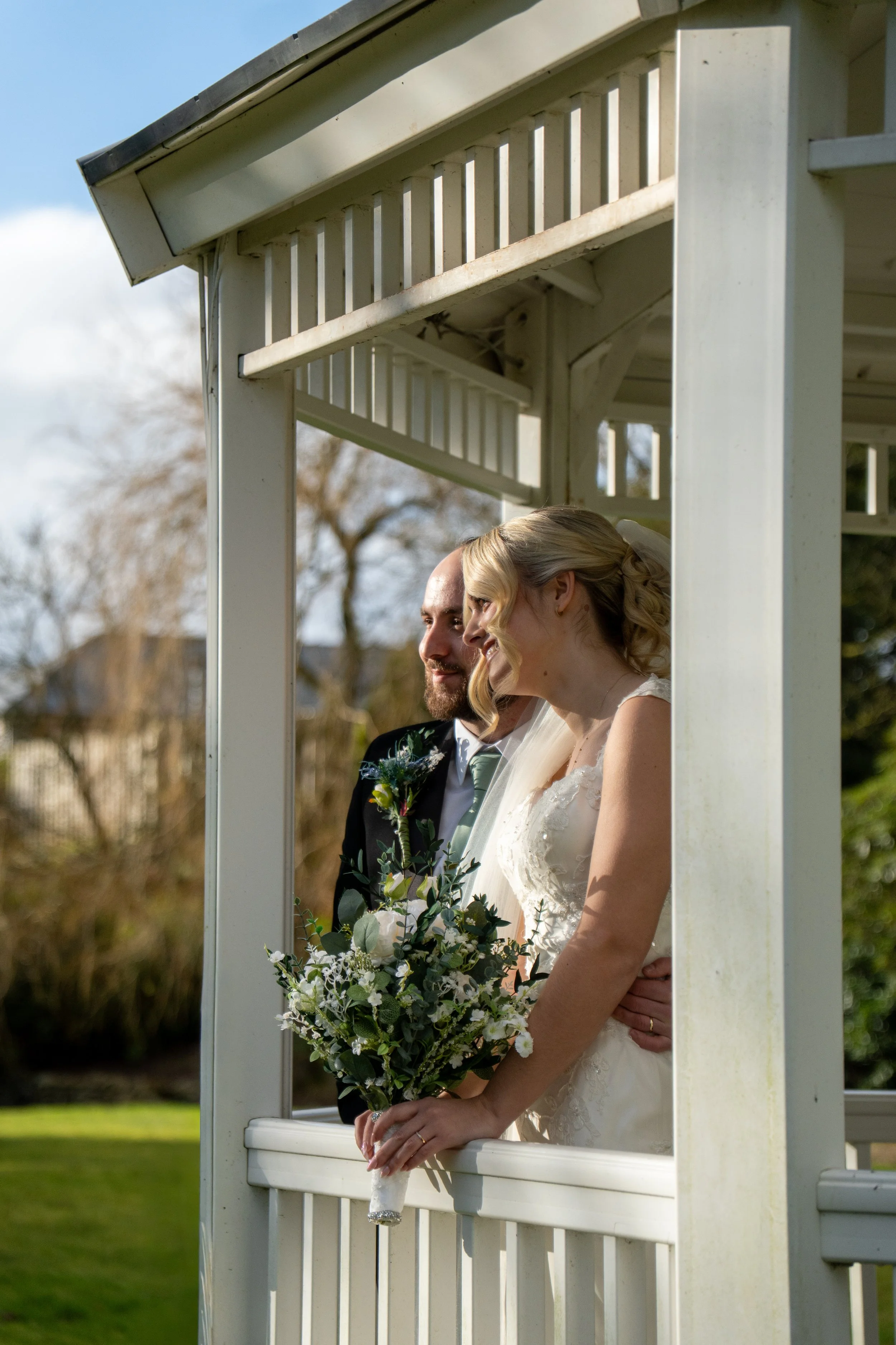 A bride and groom standing in a gazebo during an outdoor wedding ceremony, with the bride holding a bouquet of white and green flowers.