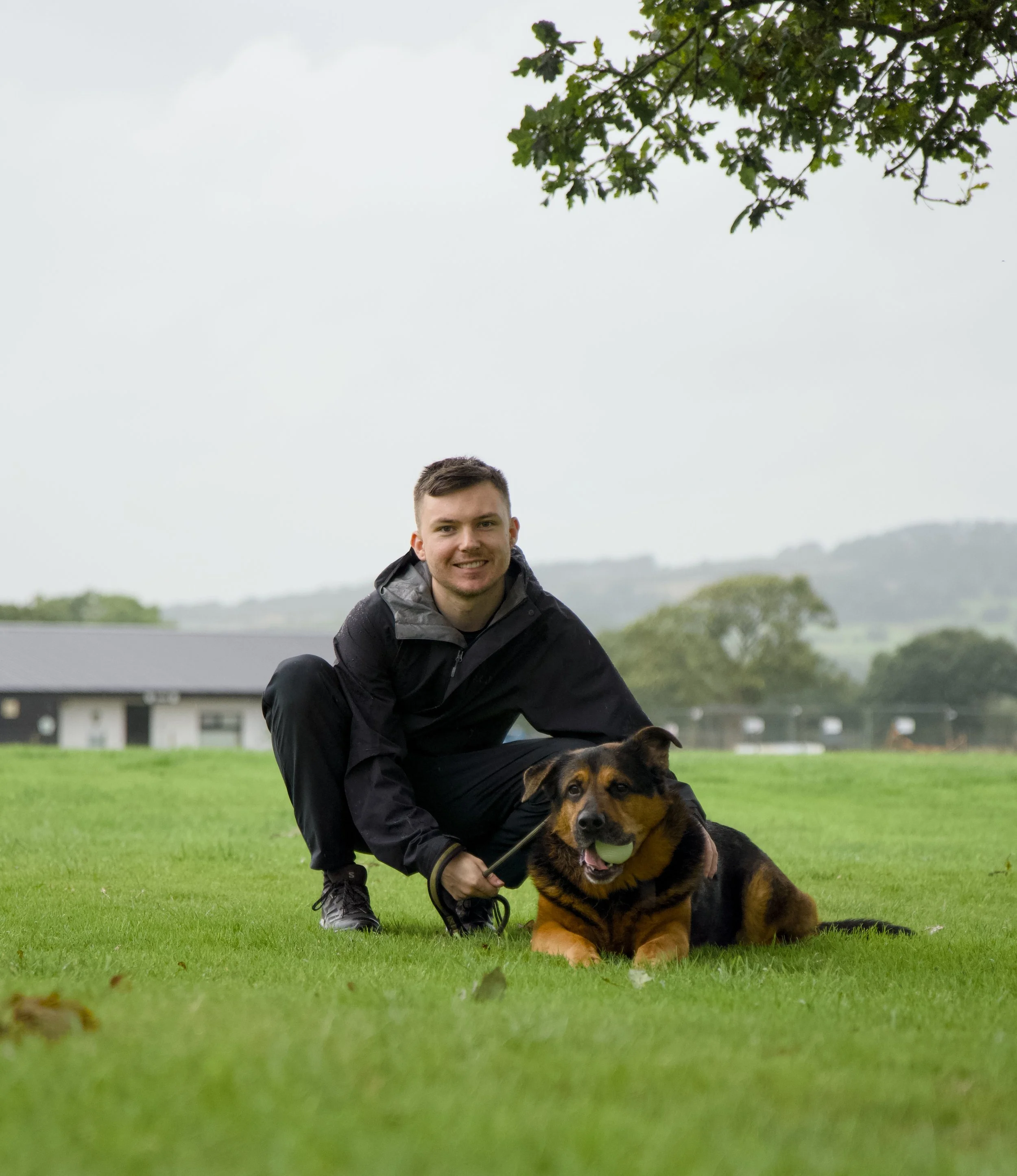 A young man with short brown hair smiling in a black jacket, crouching next to a large brown and black Rottweiler dog with a green ball in its mouth, on a grassy lawn with trees and houses in the background.