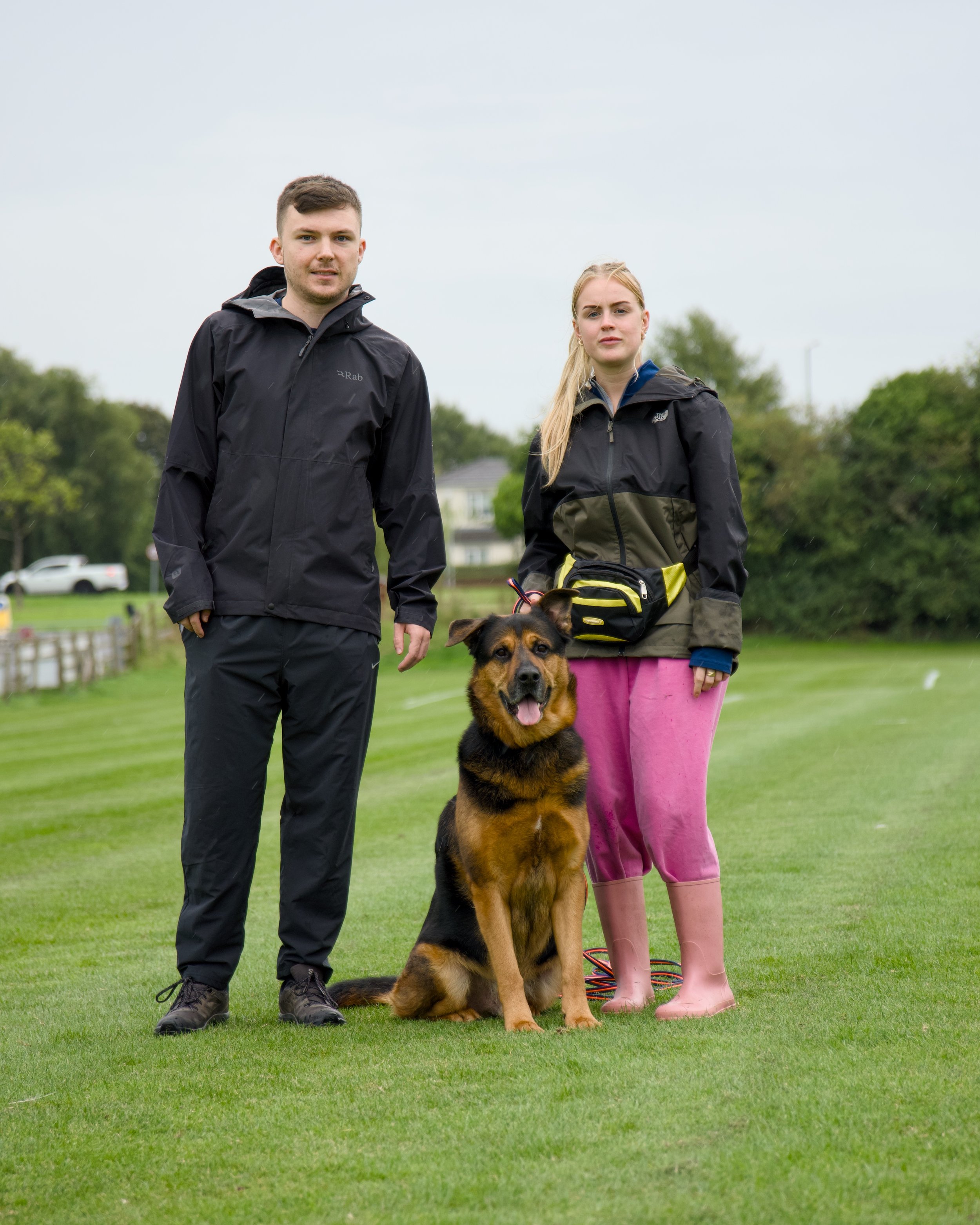 Two people and a dog standing on a grassy field on a cloudy day. The man on the left wears a black jacket, and the woman on the right wears a black and olive jacket, pink pants, and pink rain boots. The dog, a large Rottweiler, is sitting in front of the woman.