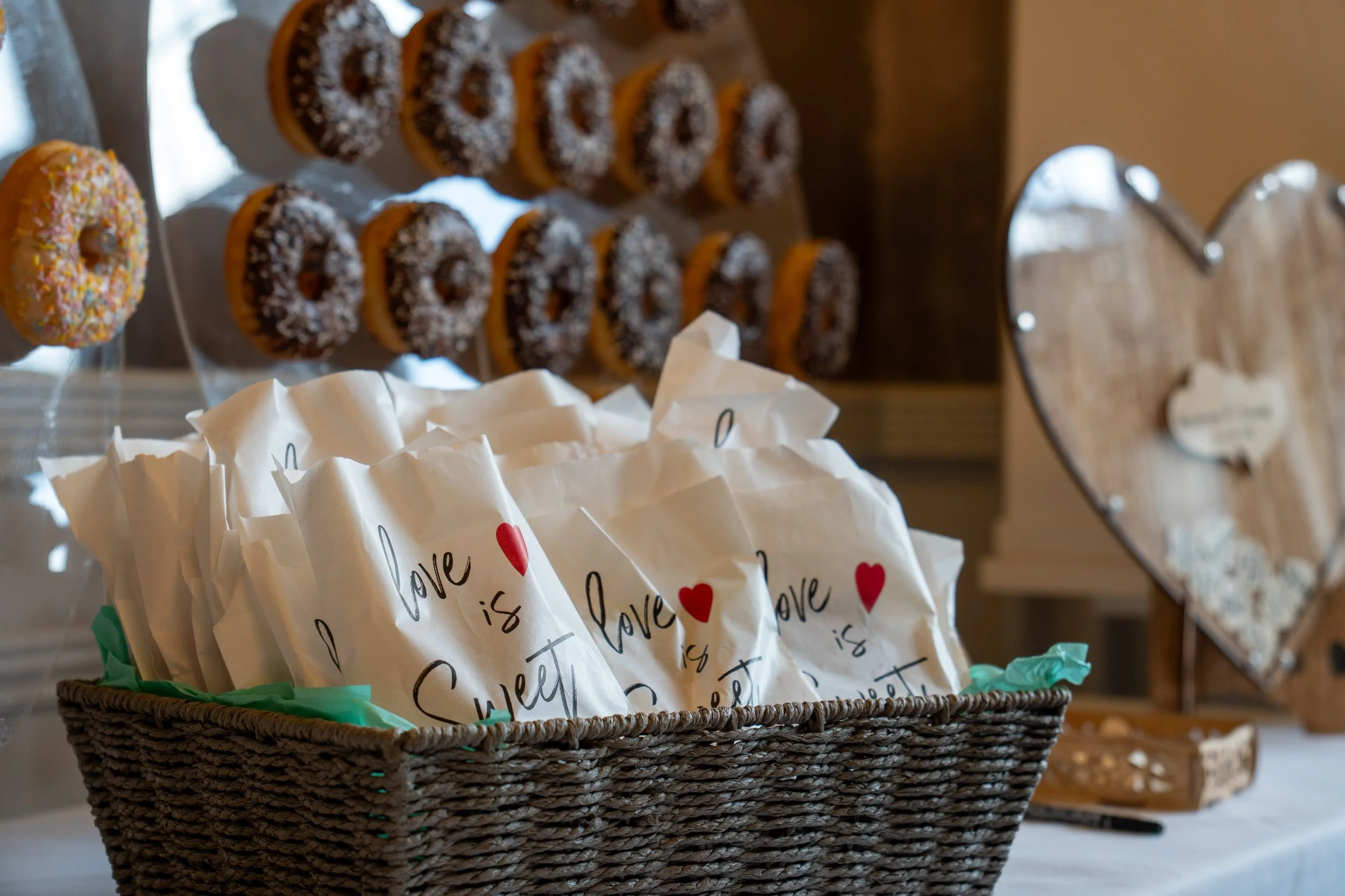 A basket filled with white paper bags decorated with red hearts and the words 'love is sweet.' Behind the basket, there are donuts with various toppings displayed on a rack and a wooden heart-shaped sign on the right.