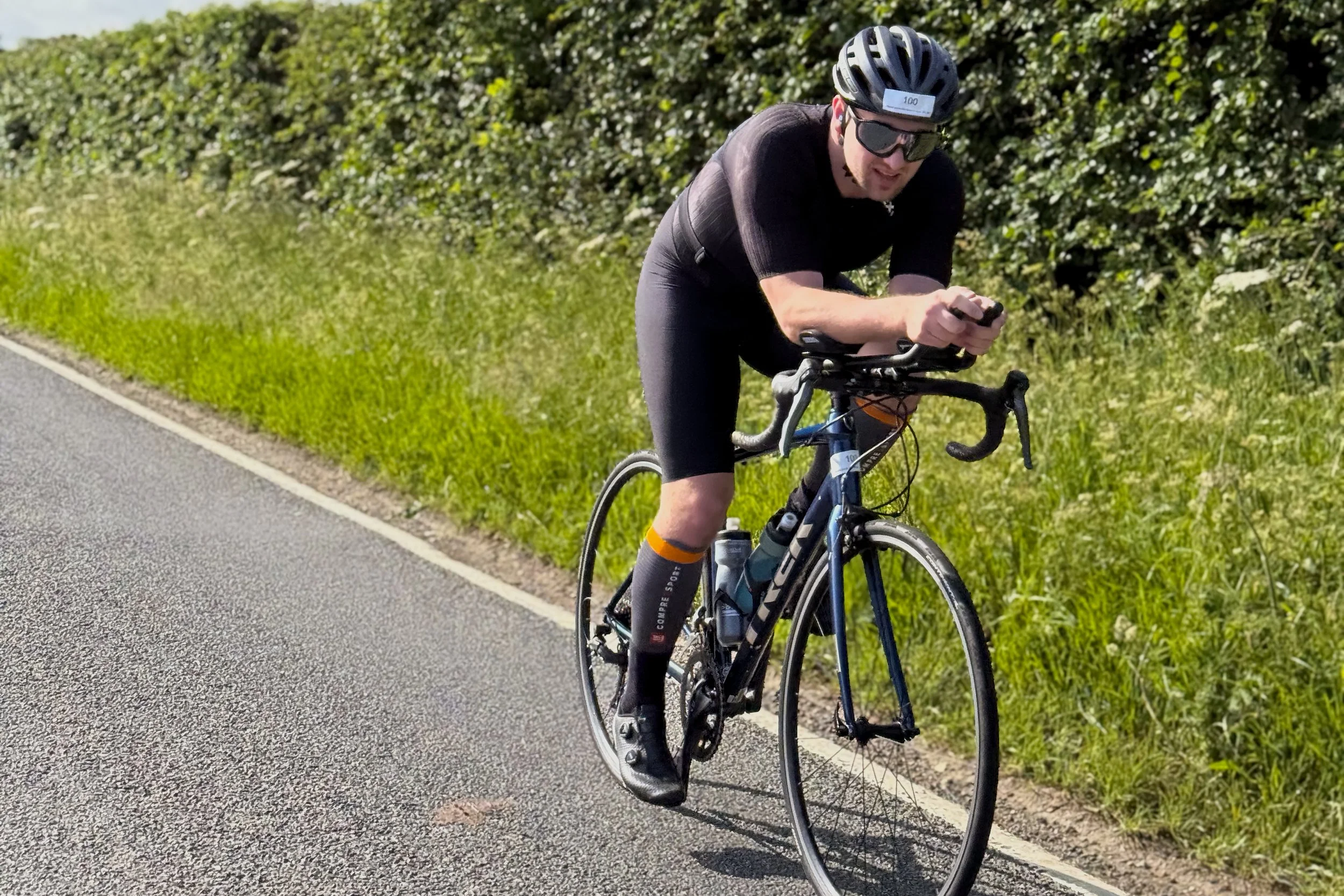 A man riding a road bike on a paved road with a green hedge on the side, wearing a black helmet, sunglasses, and cycling clothes.