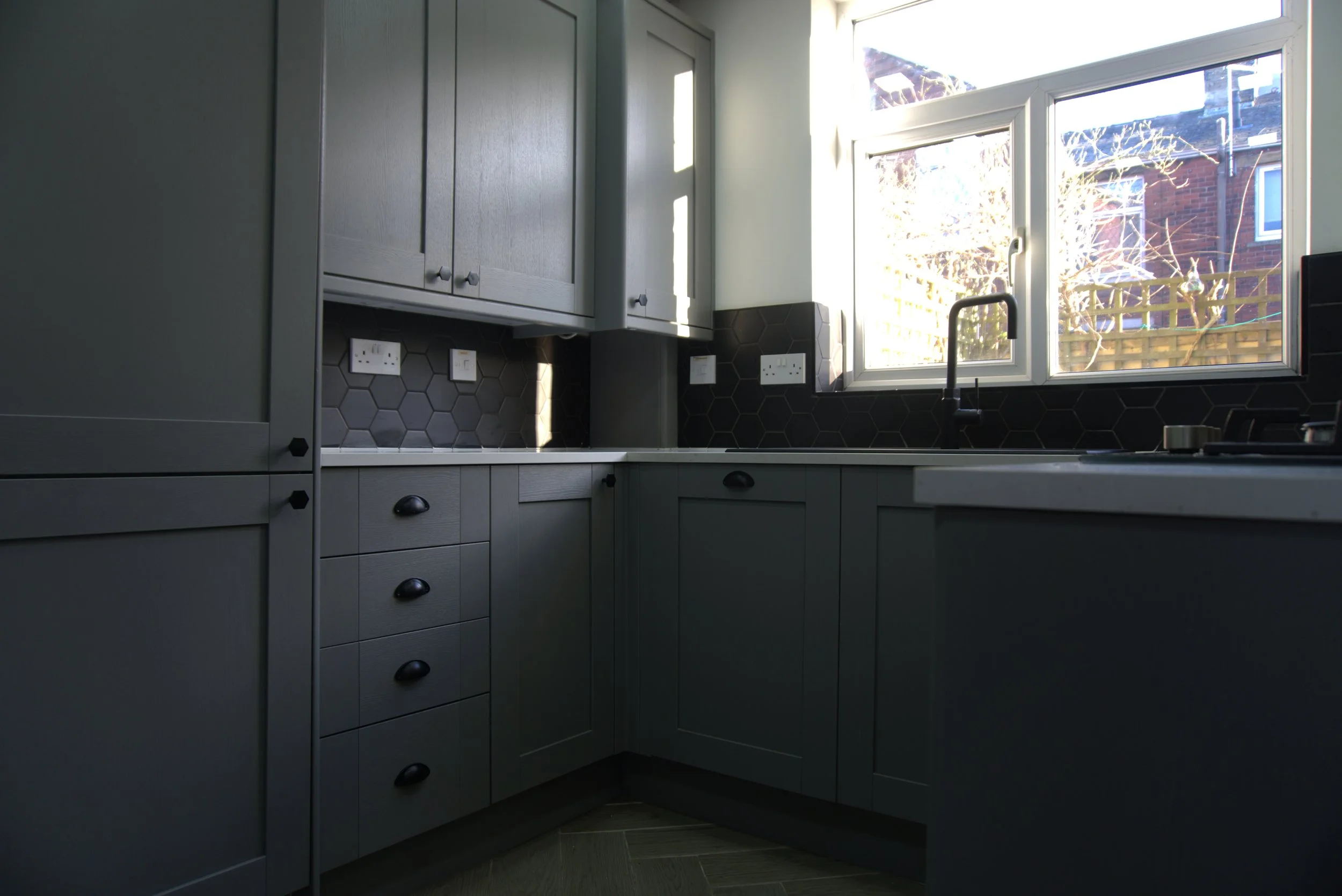 A modern kitchen with gray cabinets, a black hexagon tile backsplash, and a large window letting in natural light, located above the sink.