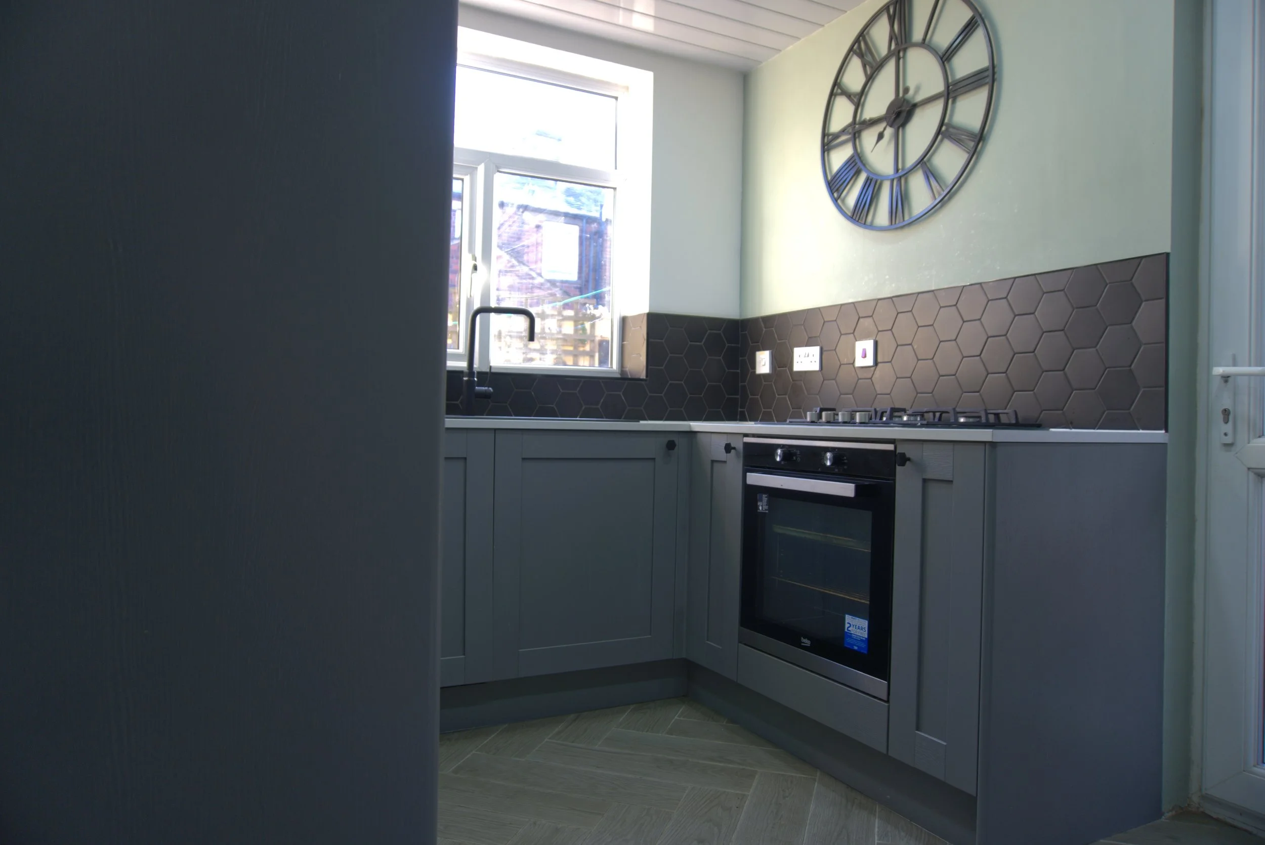 A modern kitchen with light green walls, black hexagonal tile backsplash, white cabinetry, a large black wall clock, a window letting in natural light, and built-in appliances.