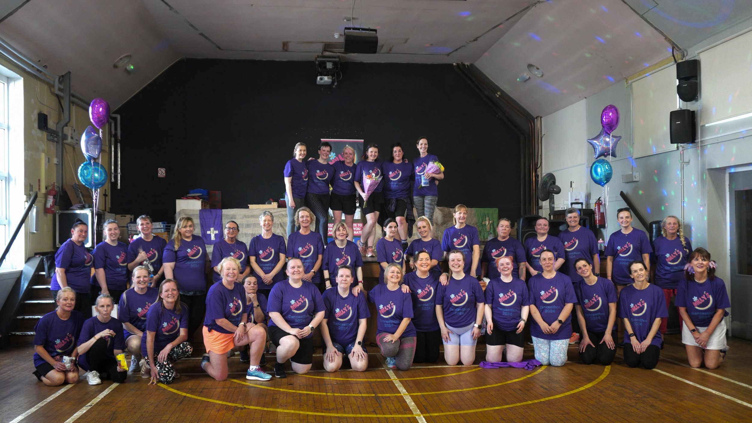 A large group of women and girls dressed in matching purple shirts gathered on stage and the floor in a community hall for a group photo, with some holding bouquets and others smiling for the camera, decorated with balloons on either side.