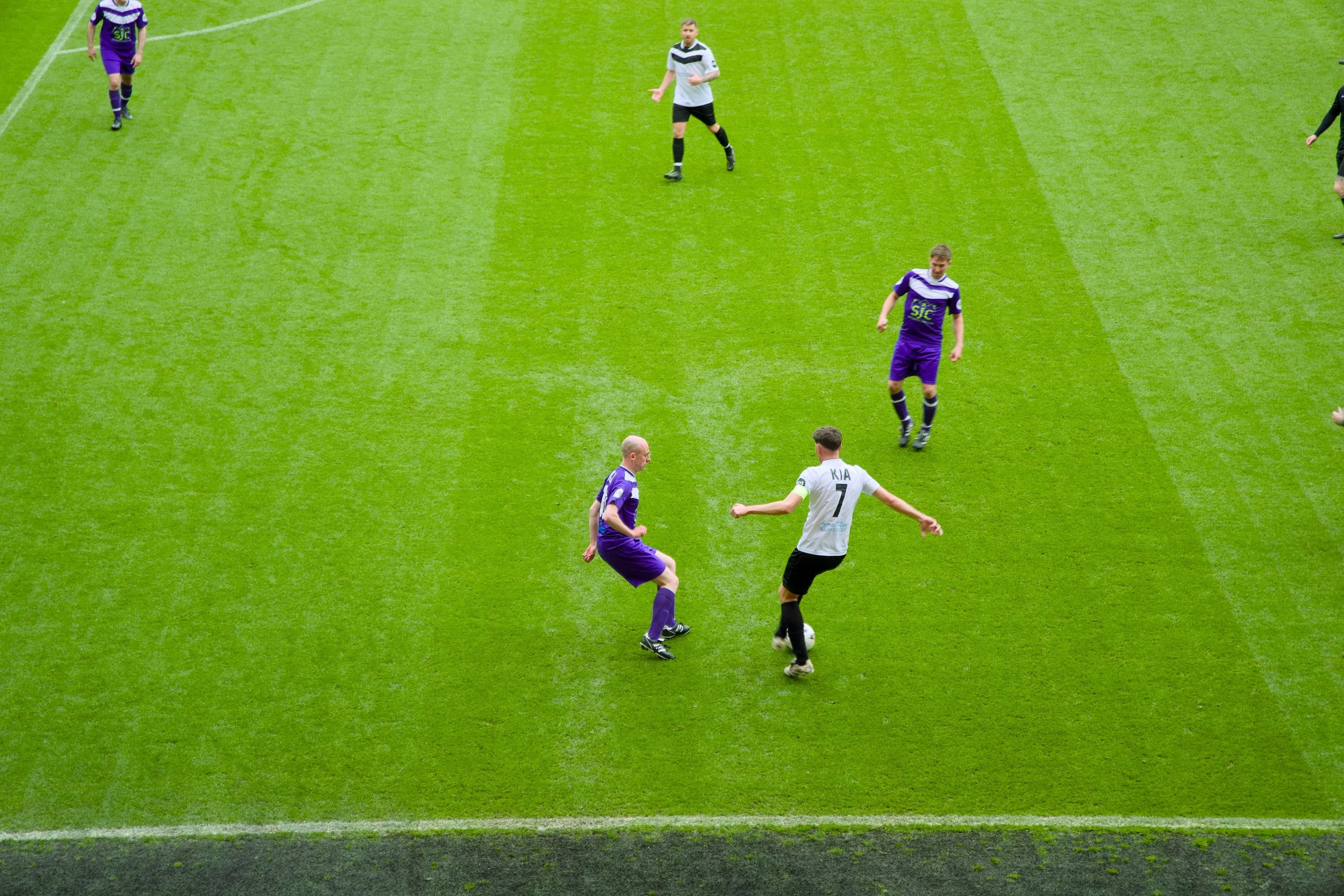 Soccer players on a green field, one in purple kit with the number 7, another in white kit with the name Kia, involved in a game, with a referee nearby.