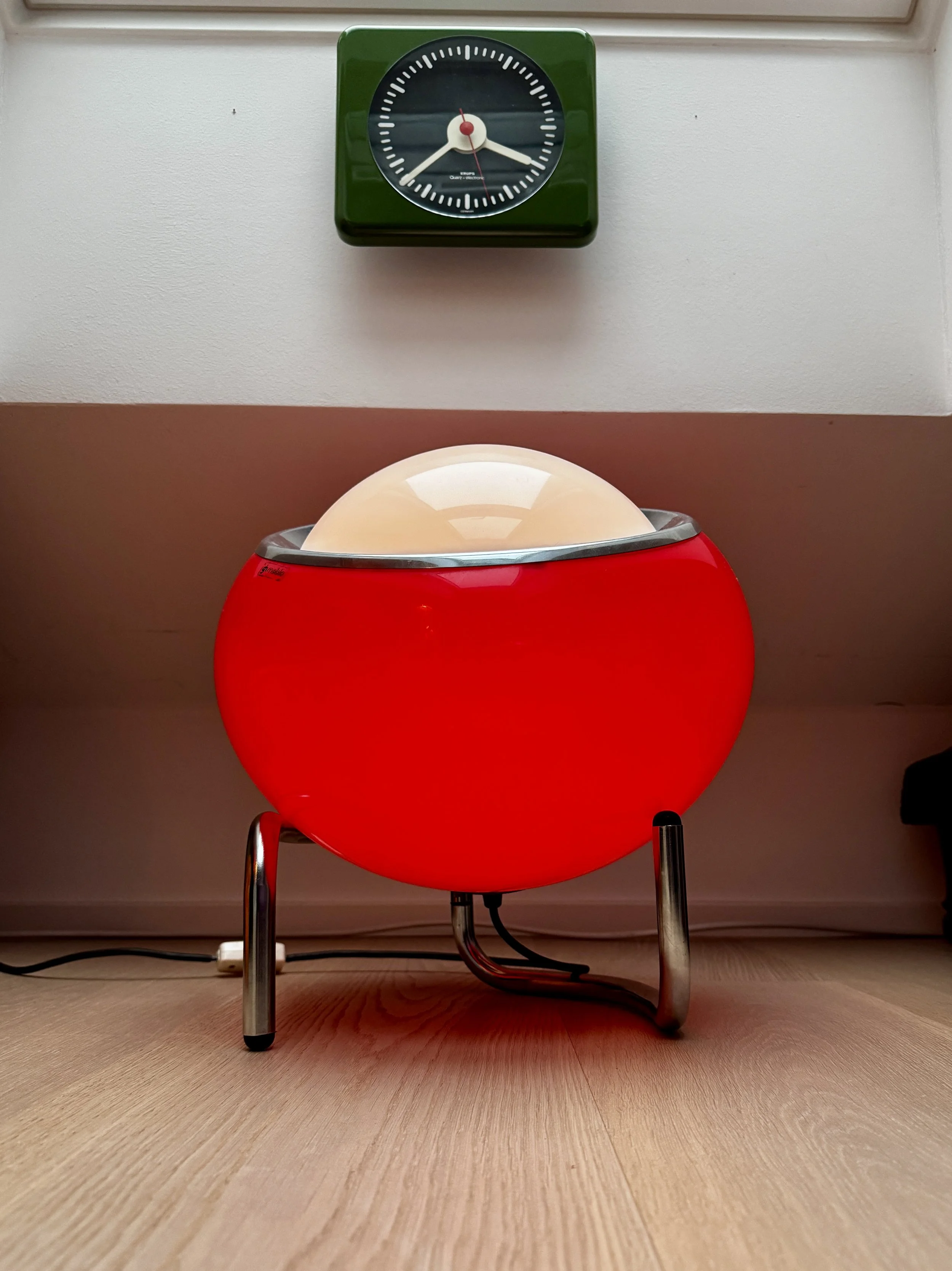 A vintage lava lamp with a red base and a glowing white globe on top, positioned on a wooden floor in front of a wall with a green clock showing approximately 3:24.