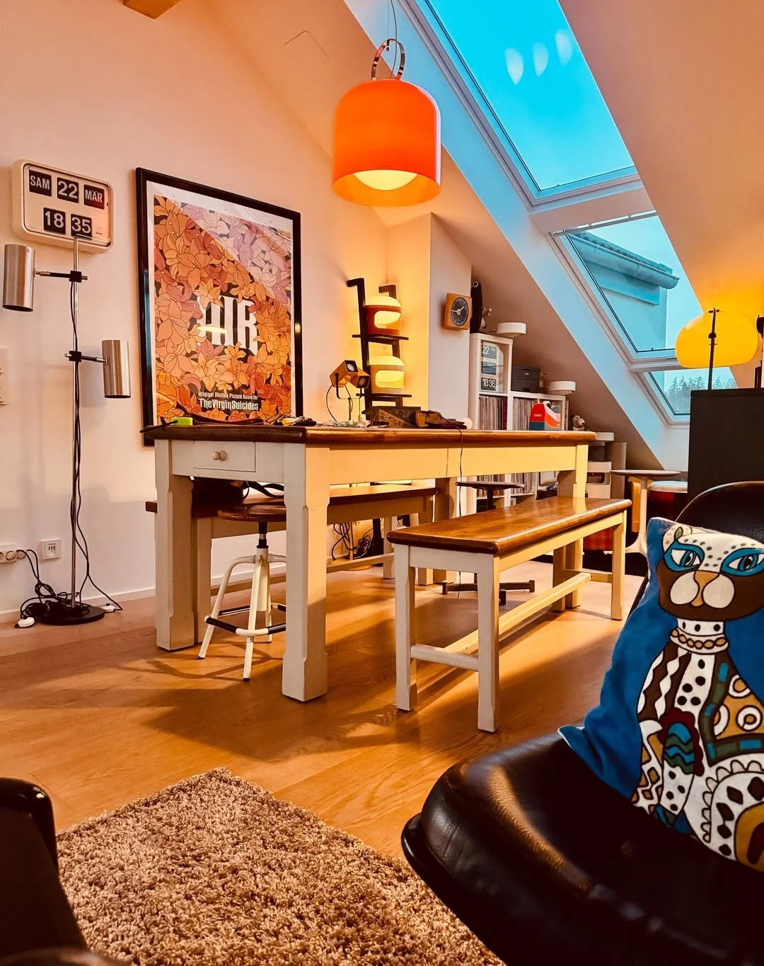 Living room with a sloped ceiling and skylight windows, containing a wooden dining table with a bench and chairs, a black leather chair with a colorful cat pillow, colorful hanging lamps, a bookshelf, and framed artwork on the walls.
