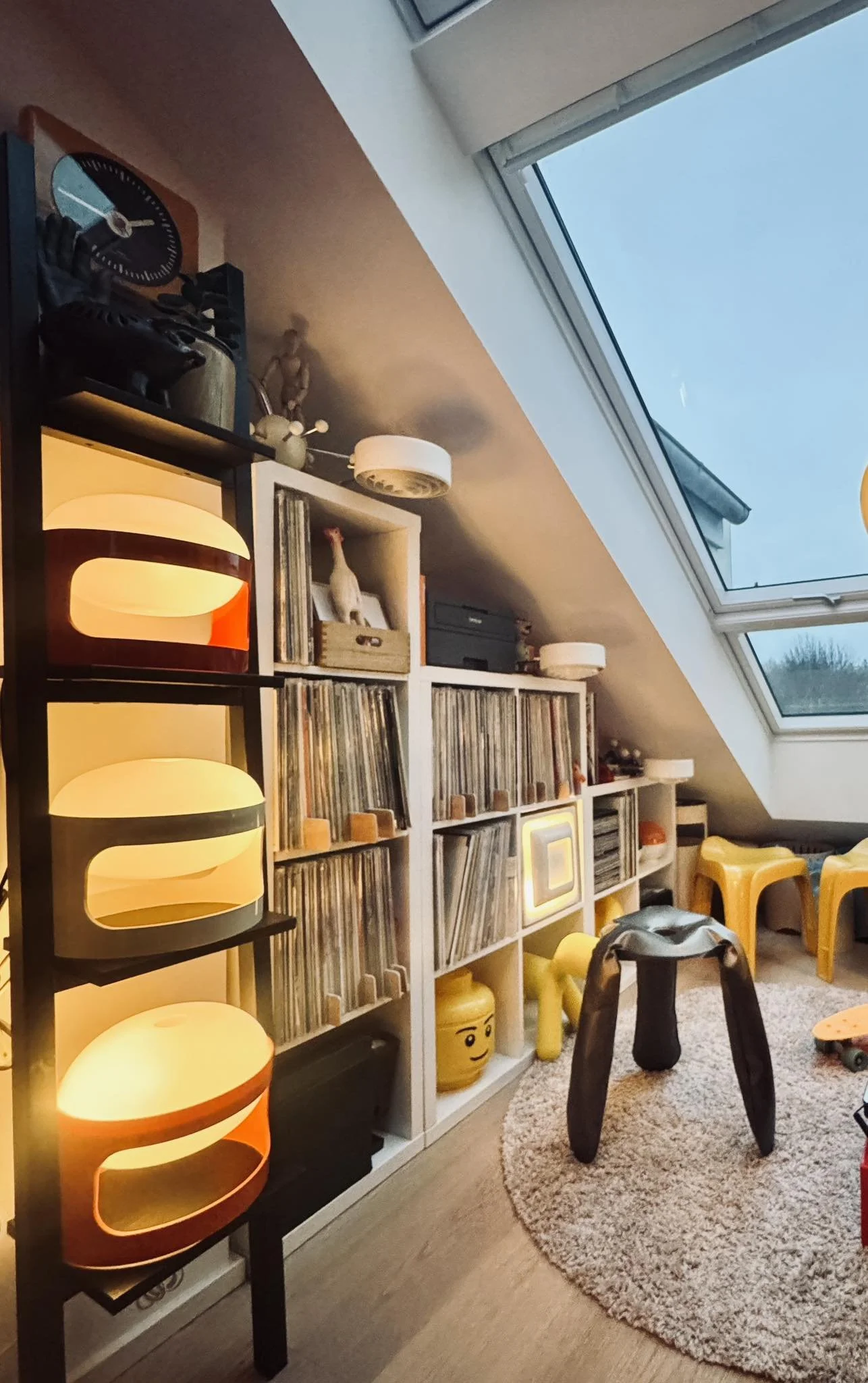 A cozy loft room with a large sloped skylight window, a white bookshelf filled with vinyl records, lamps, and decorative items, a small black table, and a yellow LEGO head-shaped storage container, with several yellow stools and a skateboard on a round beige rug.