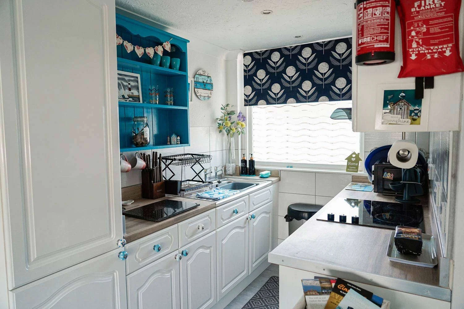 Bright kitchen with white cabinets, blue accents, and a window with patterned blinds. Contains kitchen appliances, a vase with flowers, and various kitchen items.