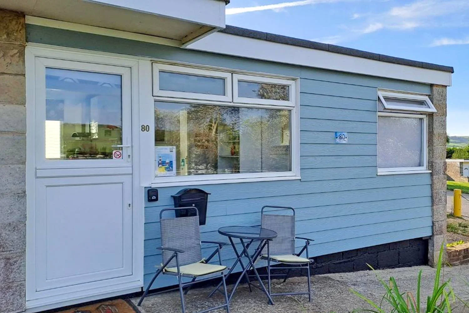 A blue house with white trim, a white door labeled '80', a large front window, and a smaller window above it. Two metal chairs with cushions and a small round table are outside, along with a mailbox and a no smoking sign.