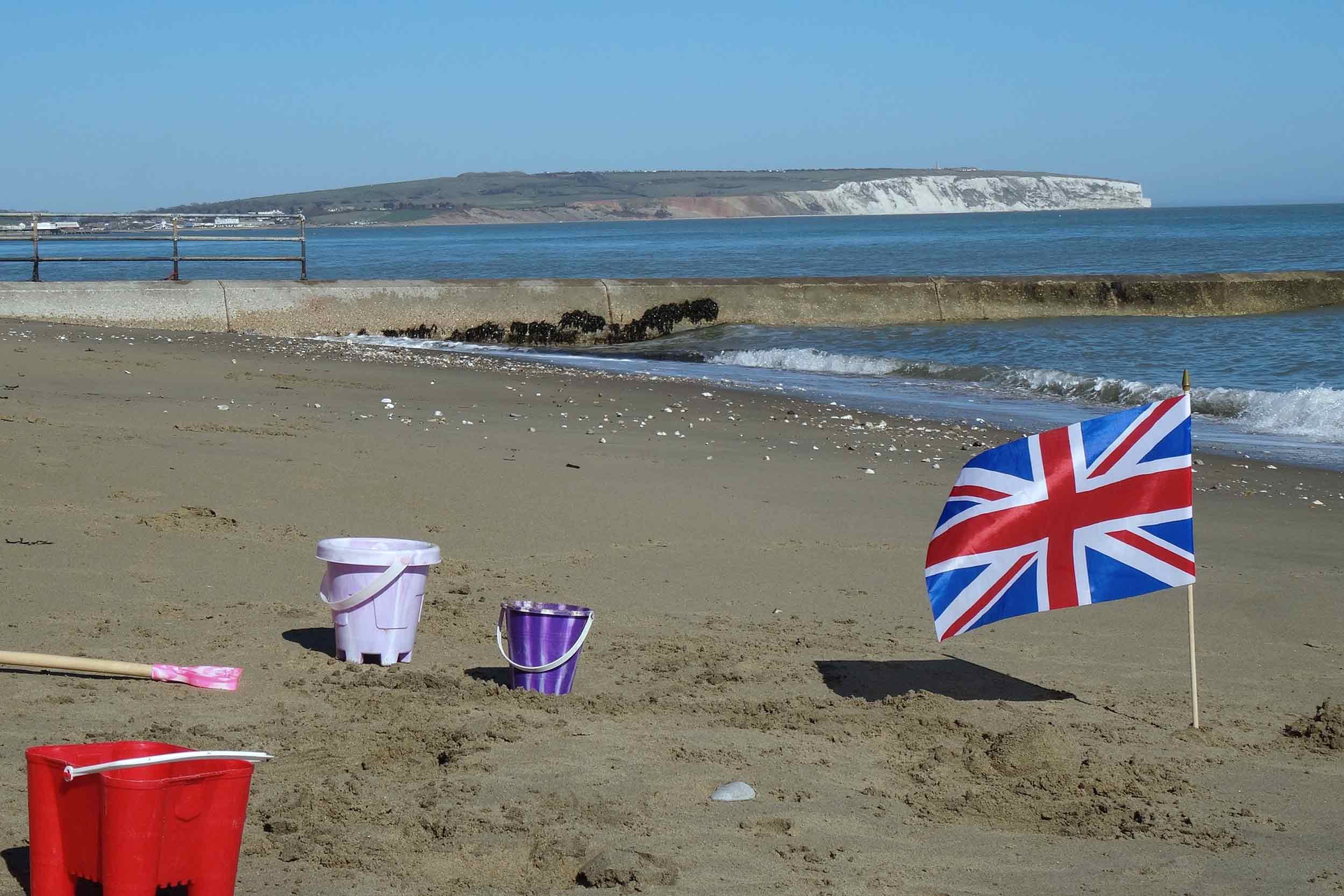 Beach scene with buckets and a Union Jack flag in the foreground, calm water, a concrete barrier, and white cliffs in the distance under a blue sky.