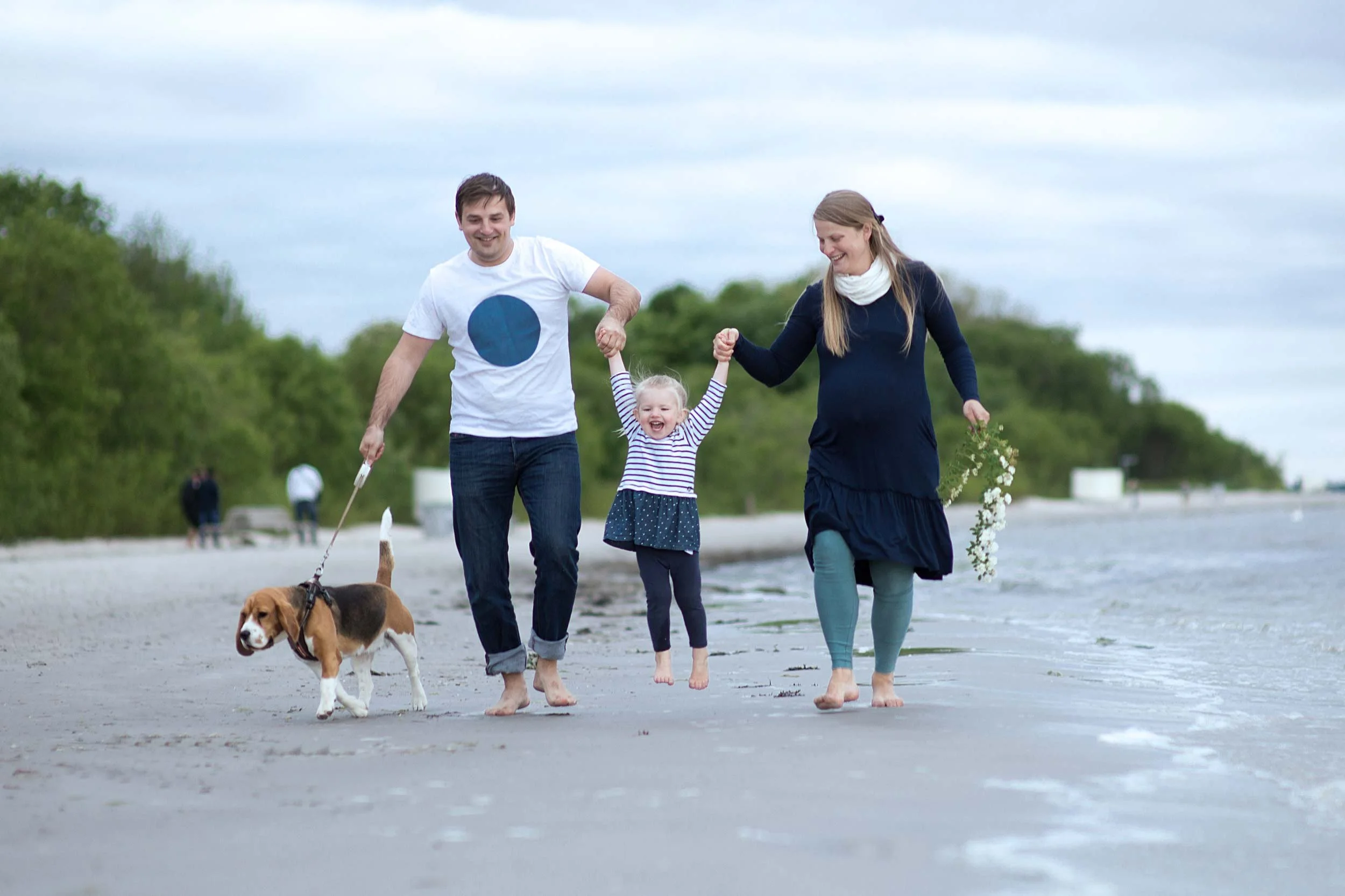 A family of three with a dog walking along the beach near the water on a cloudy day. They are smiling and holding hands, enjoying a peaceful moment together.