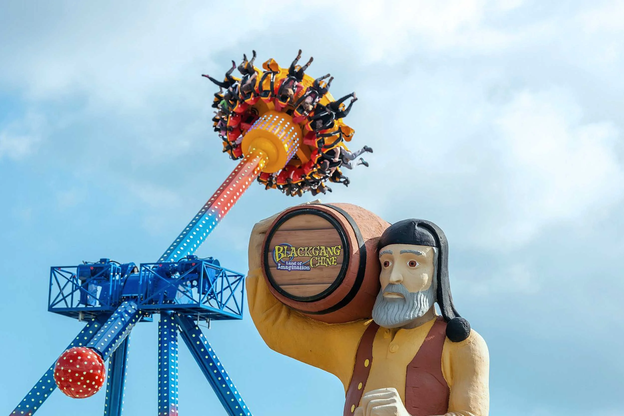 A large amusement park ride features a figure of a man with a beard, holding a large barrel labeled "Blackgang Chine". The figure appears to be in a historical or fantasy costume. In the background, there is a bright blue roller coaster with a blue track and support beams. The sky is partly cloudy.