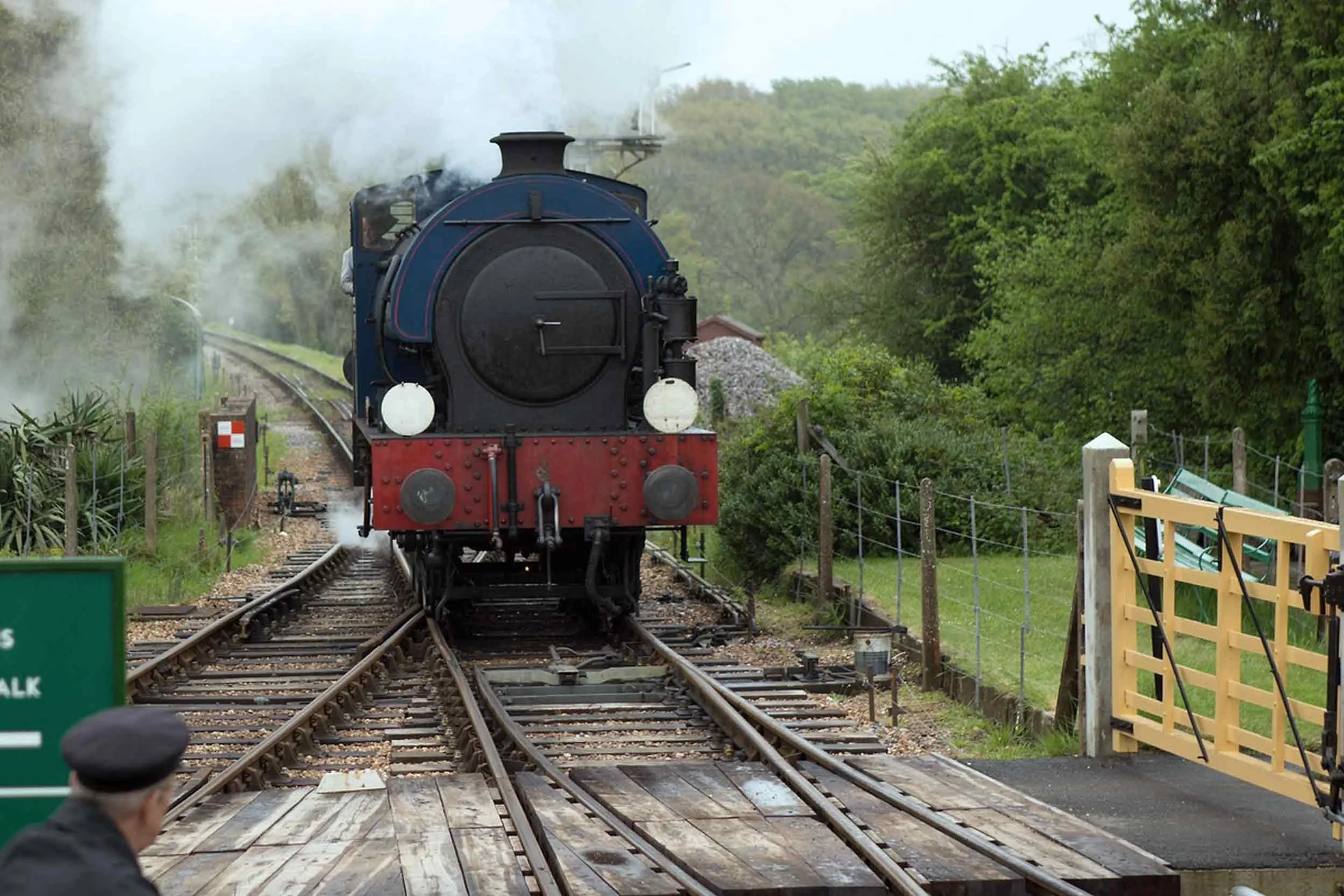 A vintage steam locomotive is moving on railroad tracks through a wooded area, emitting white steam, with fencing and benches on the side.