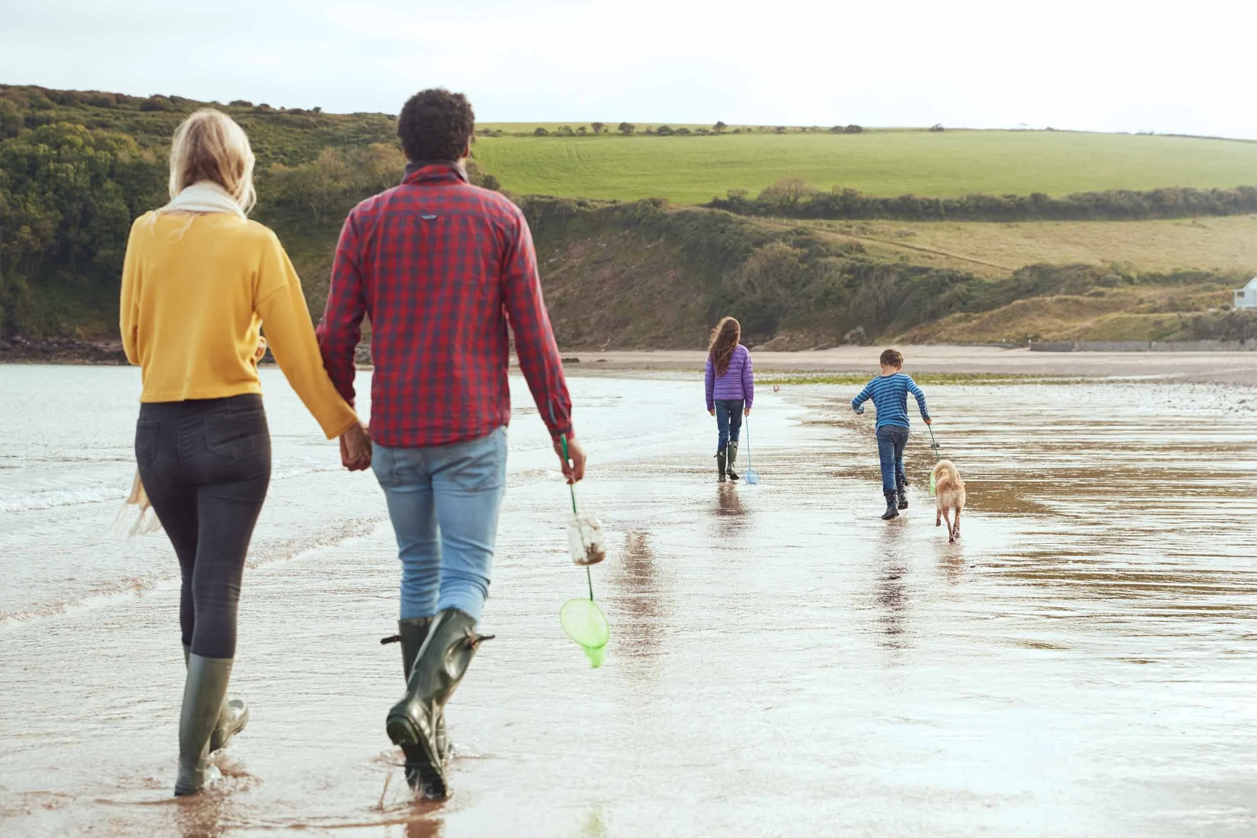 Family walking along the beach with children, some with dogs, during daytime with green hills in the background.