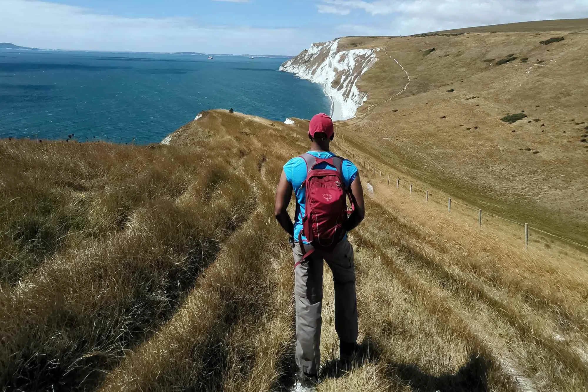 A person wearing a pink cap, blue shirt, beige pants, and a red backpack hiking along a grassy trail overlooking white chalk cliffs and the ocean in a scenic coastal landscape.