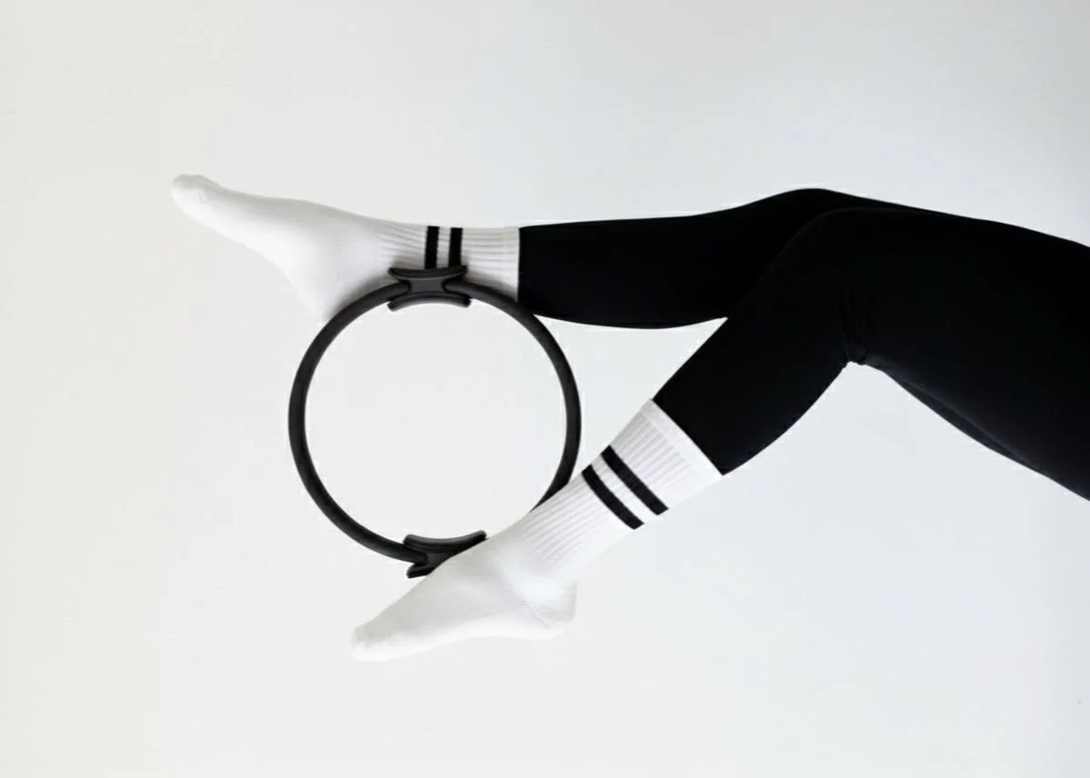 Une paire de jambes en collants noirs et chaussettes blanches avec des rayures noires, tenant une bande de levier de gymnastique en forme de cercle sur un fond blanc.