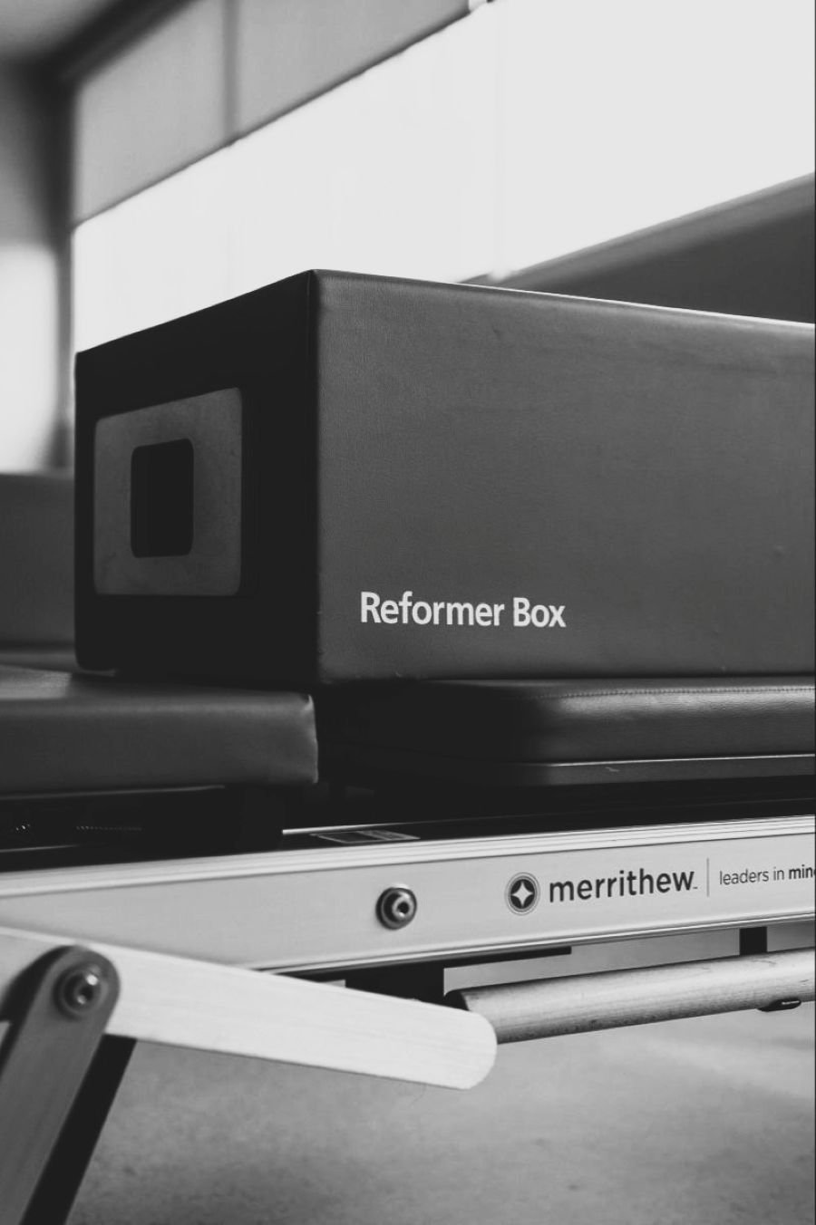 Une boîte rectangulaire de couleur sombre avec l'inscription 'Reformer Box' sur le côté, placée sur une surface métallique avec des éléments mécaniques visibles, dans un environnement probablement de fitness ou de rééducation, en noir et blanc.