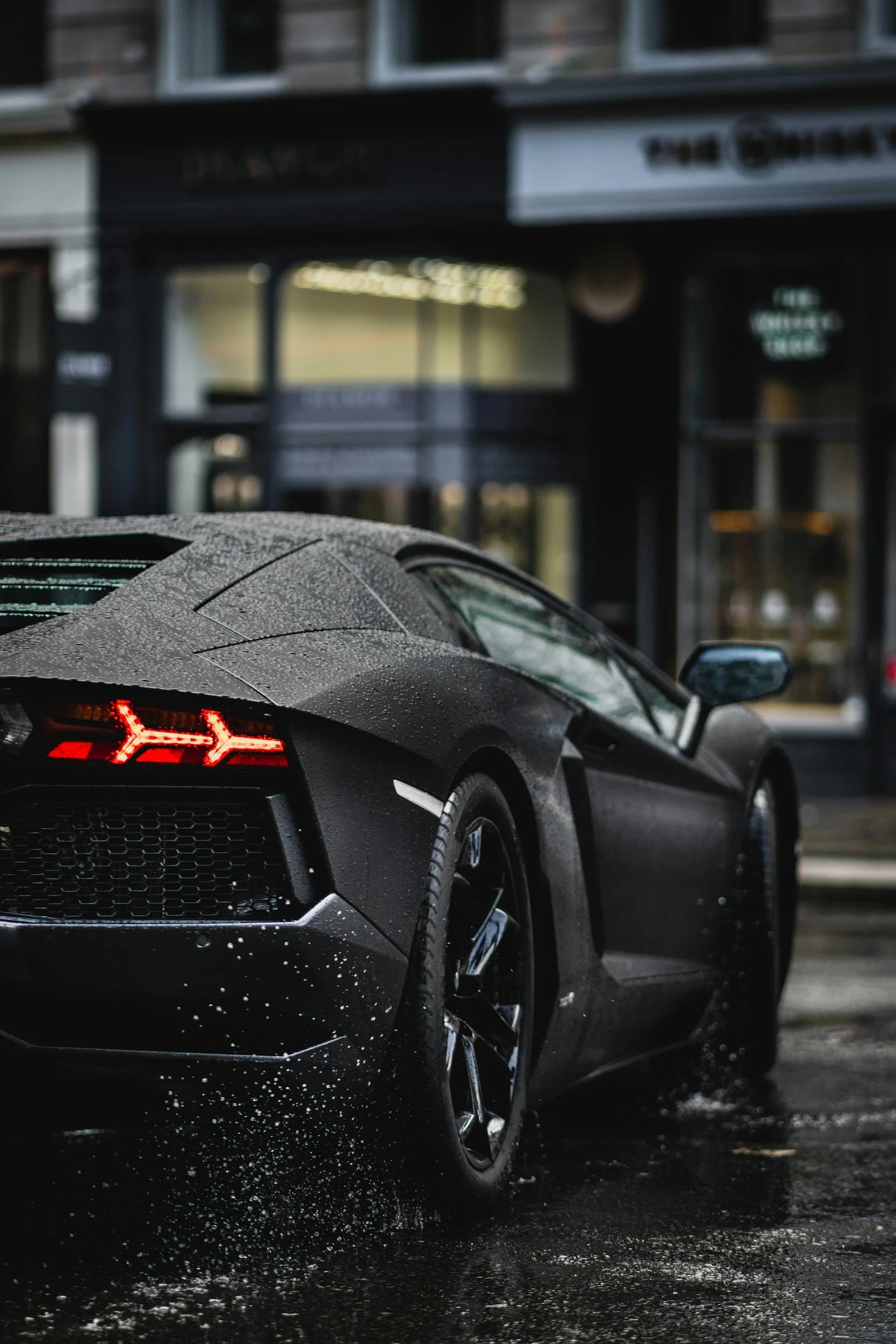 A black sports car with water droplets on its surface parked on a wet street in front of a storefront.
