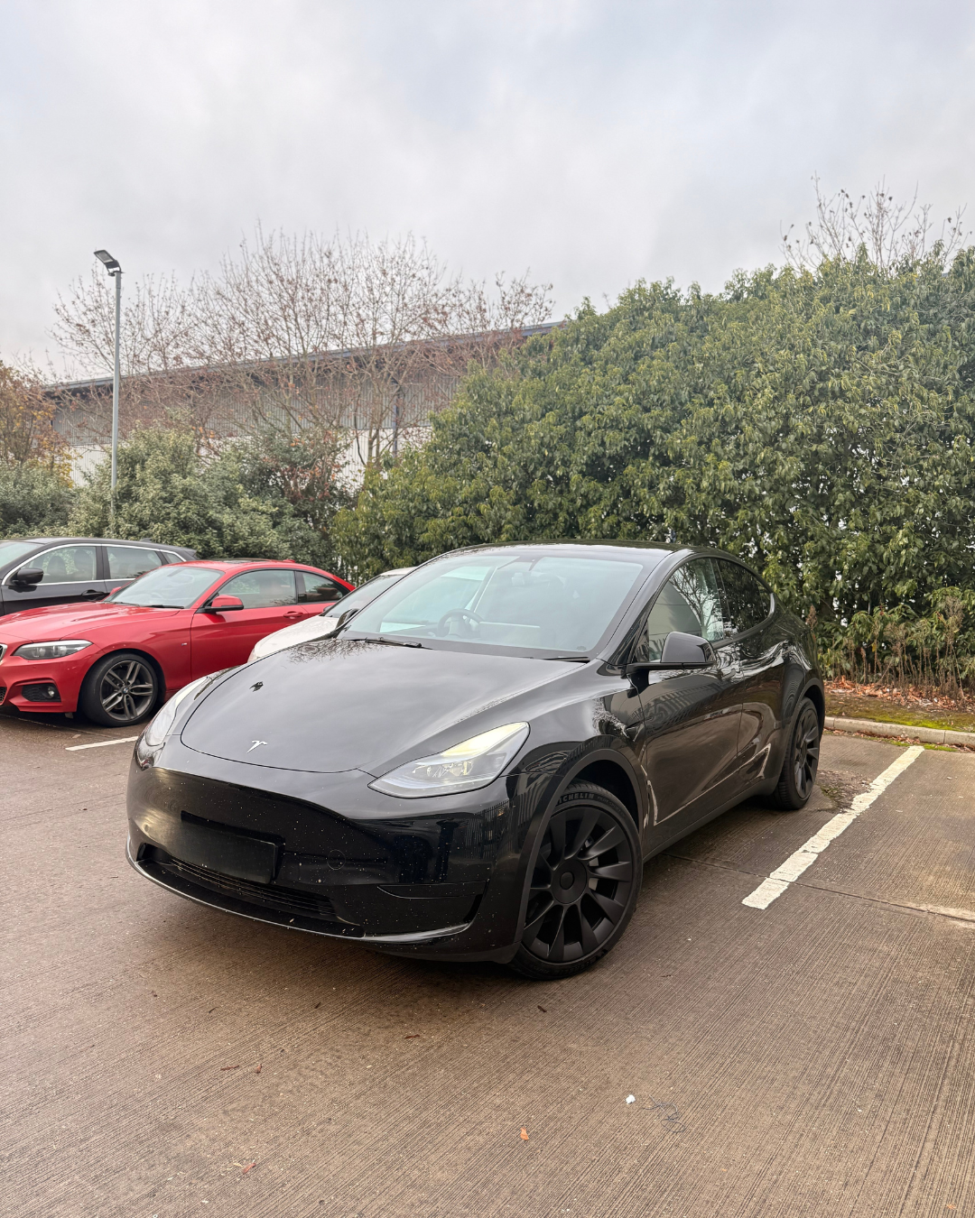 Black Tesla Model 3 parked in a parking lot next to a red sports car and another vehicle, with trees and a building in the background.