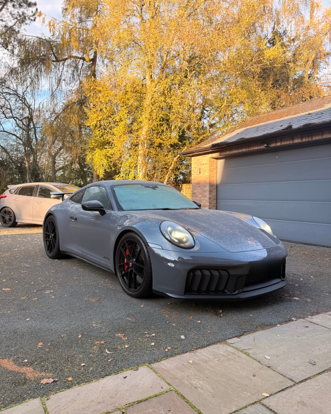 Gray sports car with black wheels and red brake calipers parked on driveway, with autumn trees and a garage in the background.