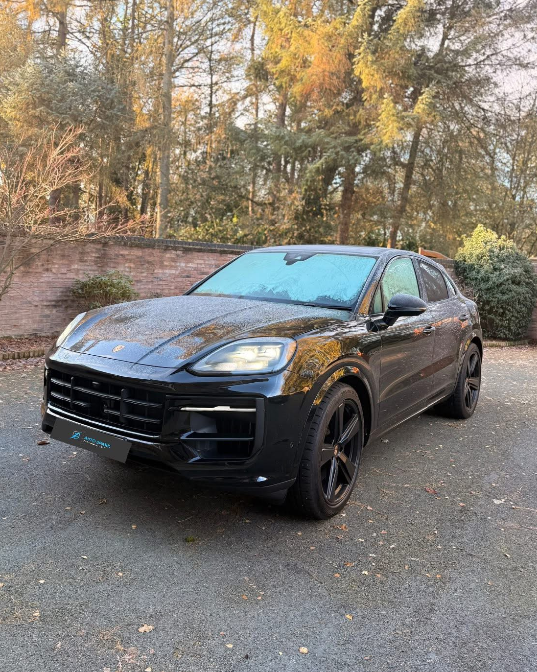 Black Porsche SUV parked in an outdoor lot with trees and a brick wall in the background, some frost on the windshield.