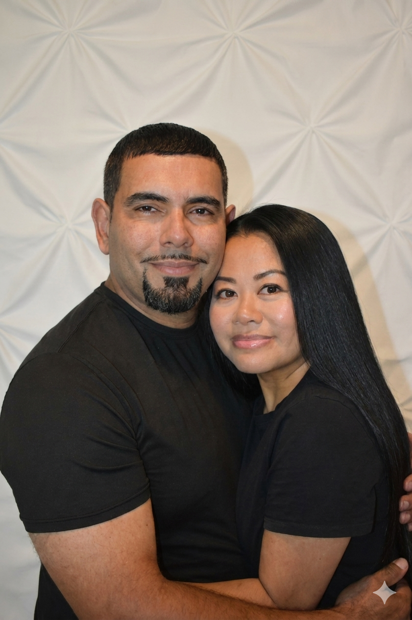 A man and woman hugging each other, both smiling, standing against a patterned white backdrop.