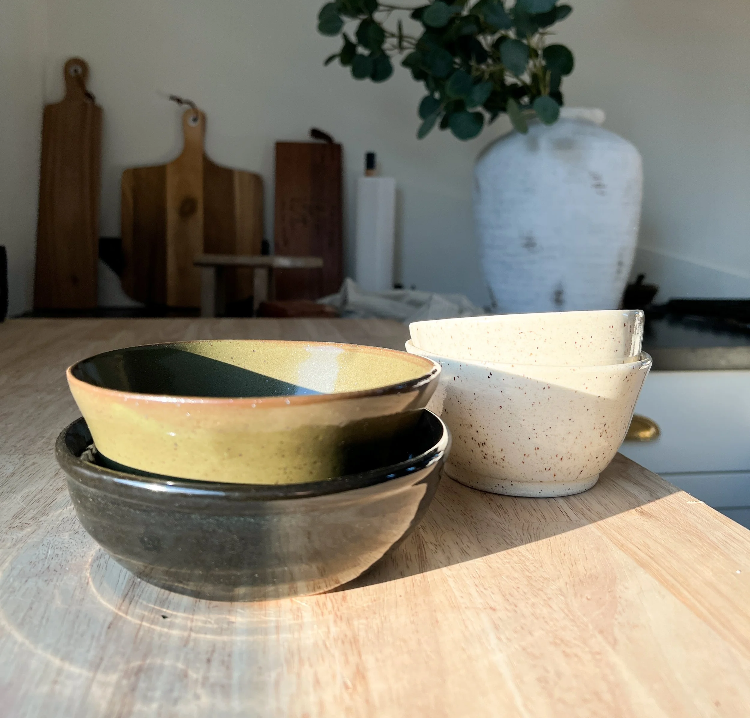 Close-up of three ceramic bowls on a wooden table with a blurry background that includes wooden cutting boards, a white vase with green leaves, and a light-colored wall.
