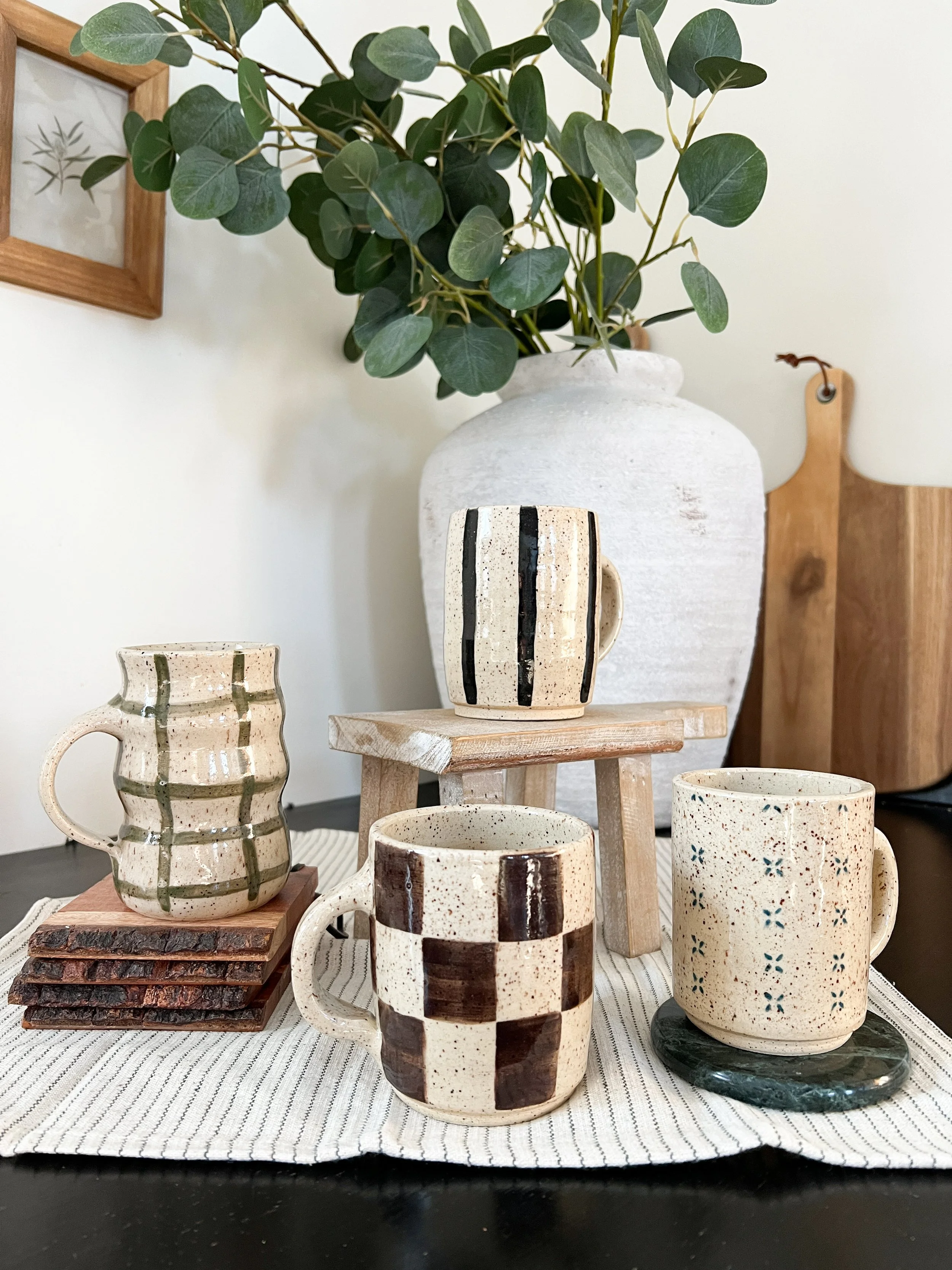 Various handmade ceramic mugs and cups with geometric and floral patterns displayed on wooden and stone pedestals on a striped cloth tablecloth.
