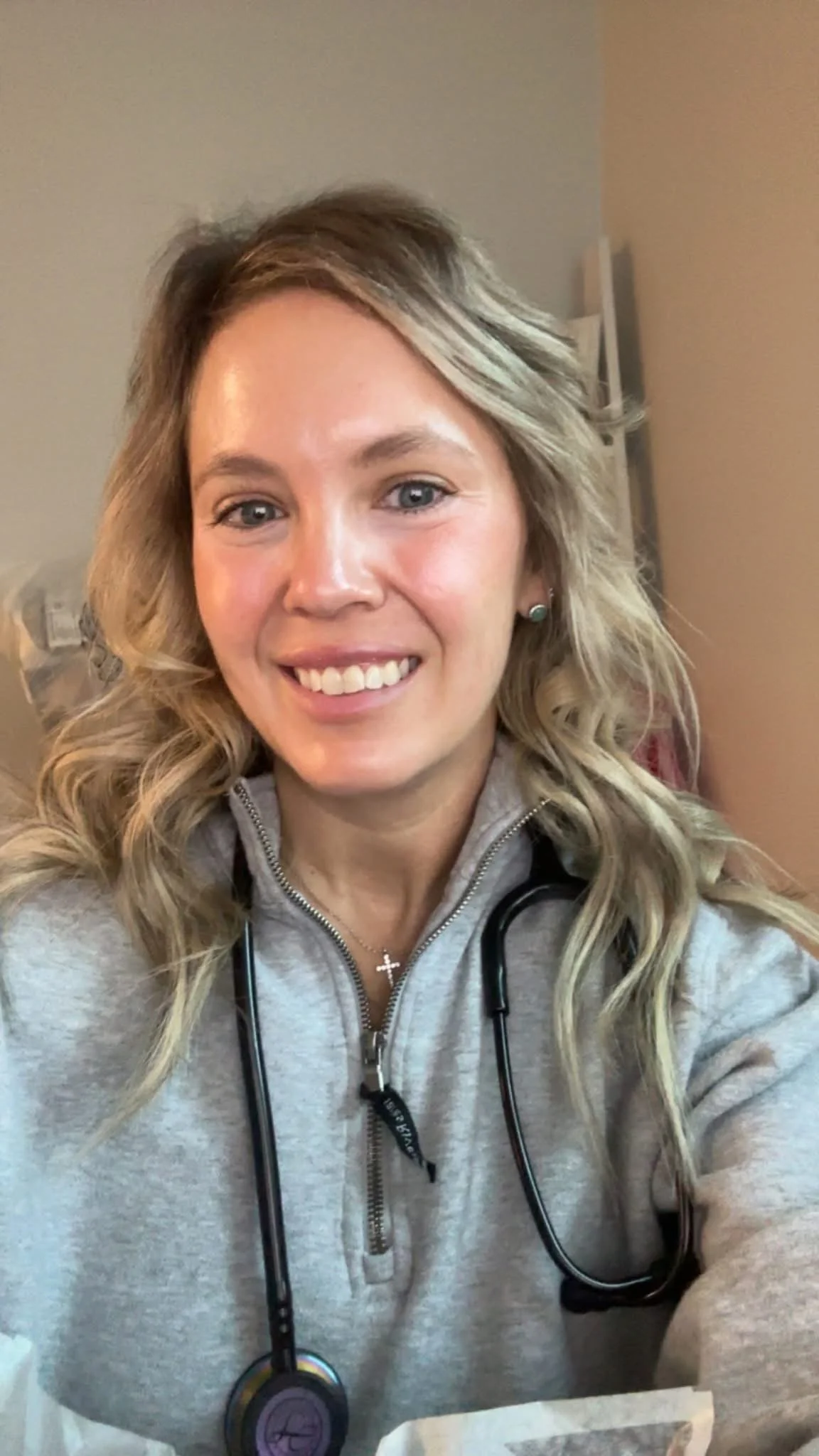 A woman with blonde, wavy hair smiling at the camera, wearing a gray zip-up jacket and a stethoscope around her neck.