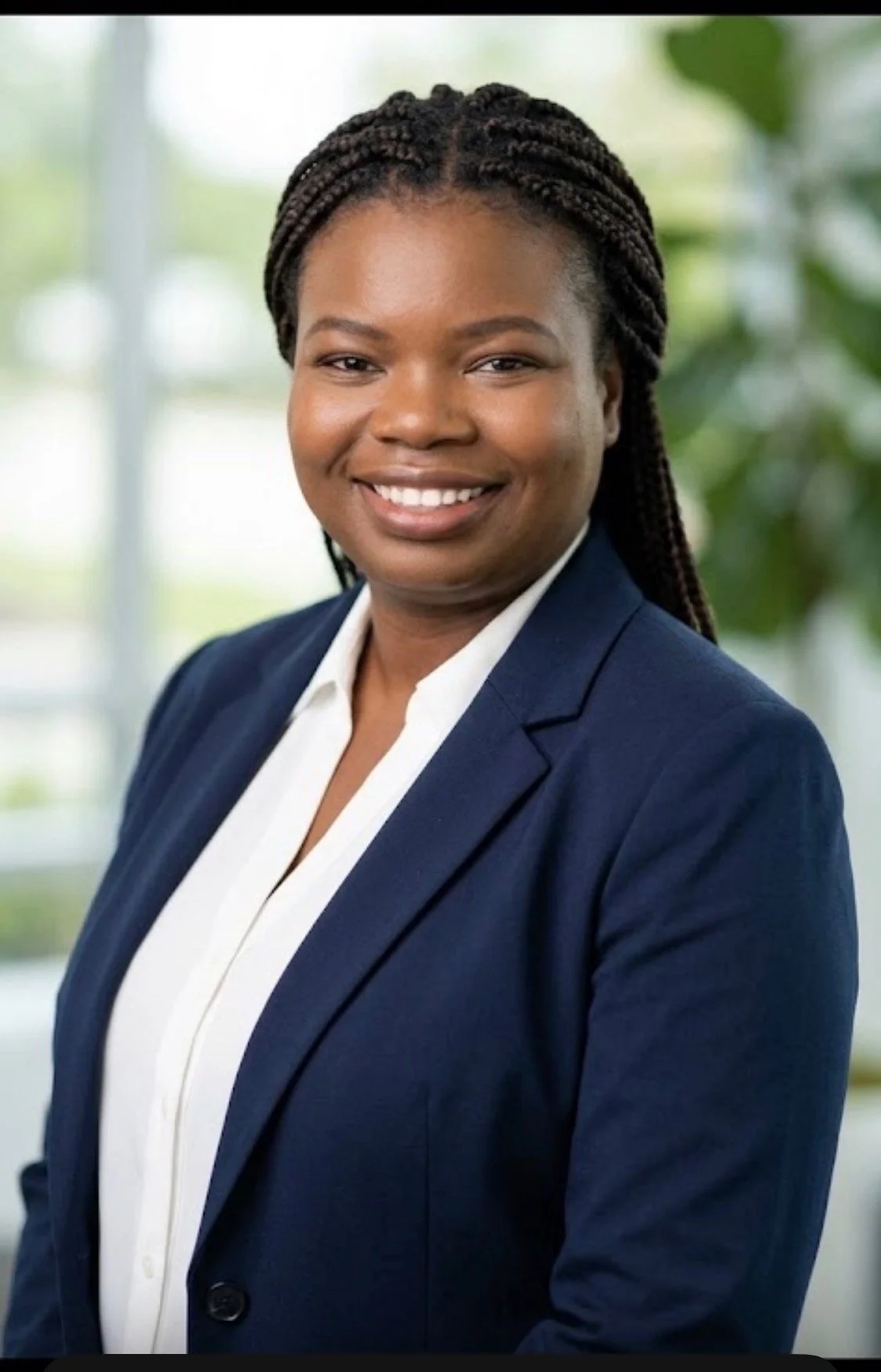 A professional woman with braided hair, wearing a navy blazer and white shirt, smiling in an office setting with blurred green plants in the background.