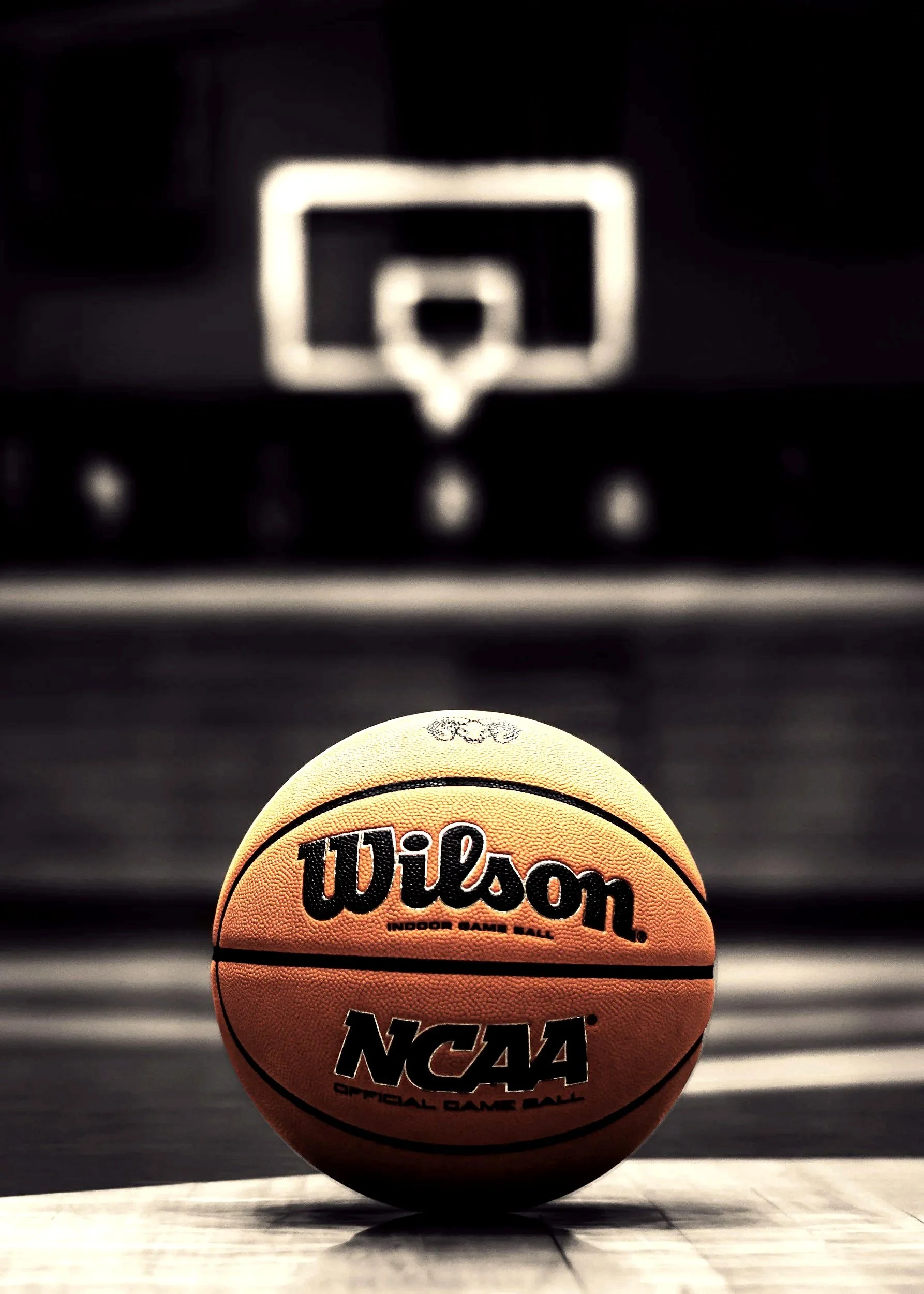 A Wilson NCAA official game basketball on a court, with a blurred basketball hoop in the background.