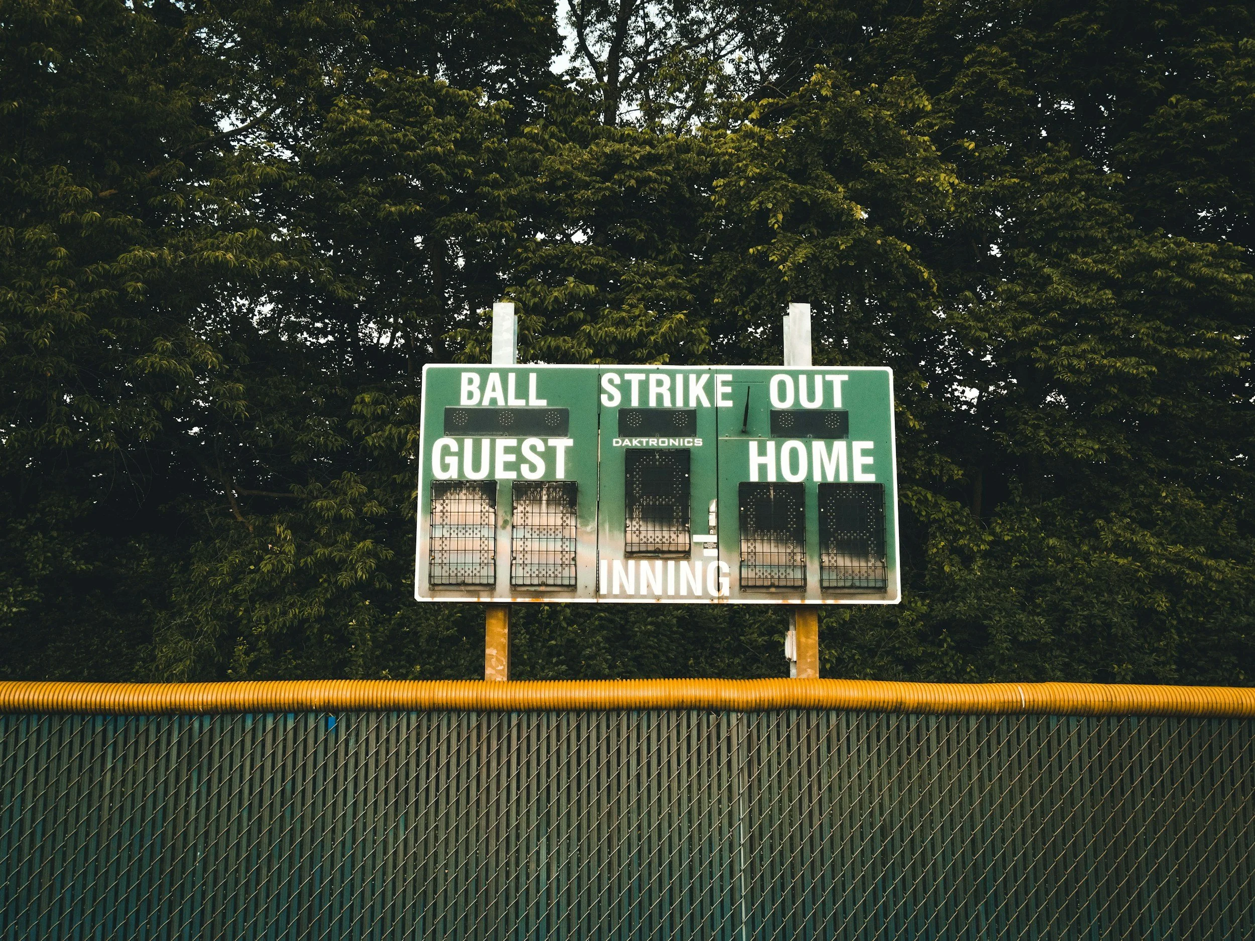 Scoreboard at a baseball field showing no current scores, with a green backdrop of trees and a metal fence in the foreground.