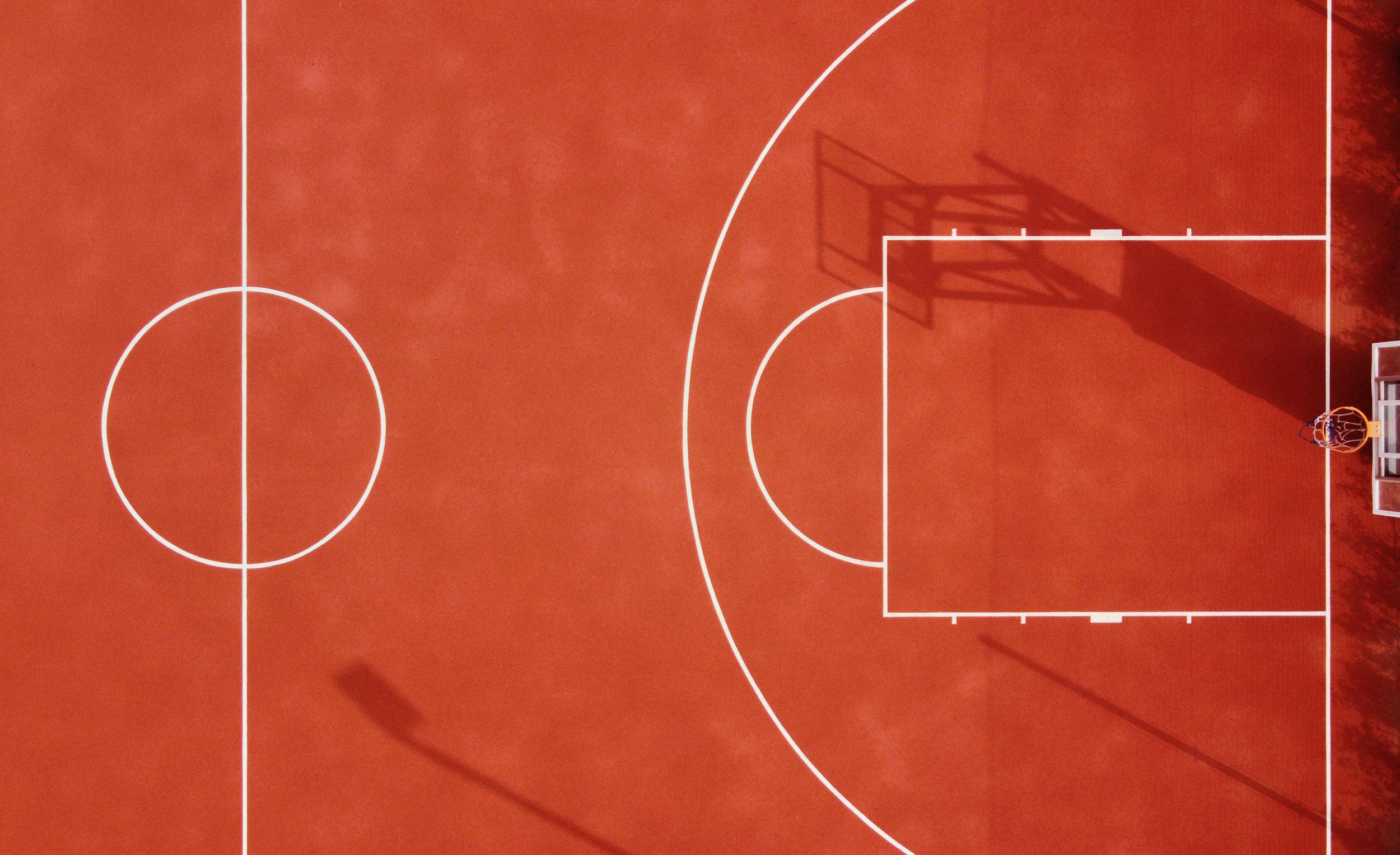 An overhead view of a basketball court with a bright orange surface, white lines, a basketball hoop with a backboard, and a basketball net. The court's shadow is visible on the surface.
